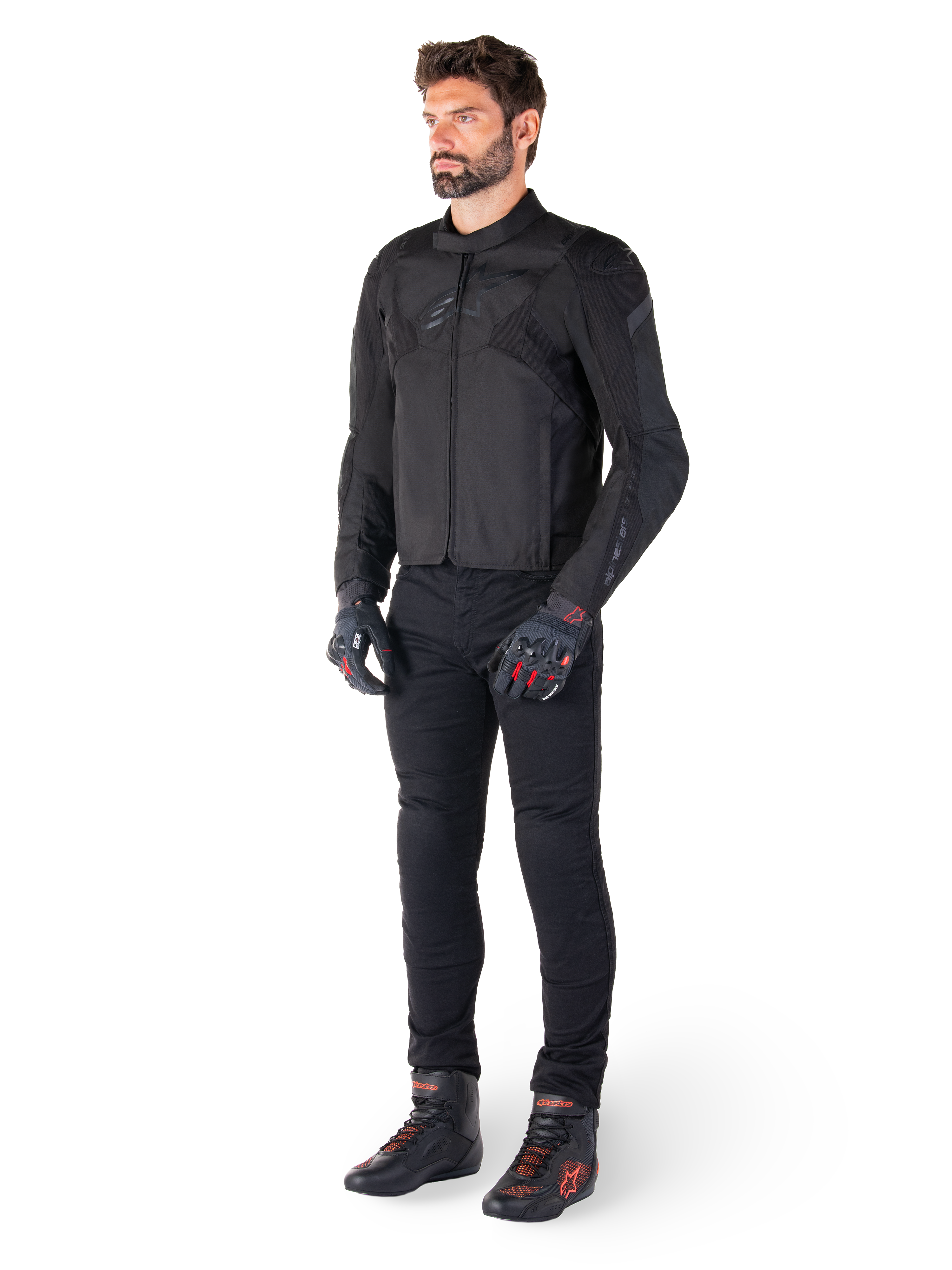 Mannelijk model draagt de Alpinestars T-Jaws V4 Waterdichte Jas in het zwart, gecombineerd met een zwarte motorbroek, zwarte handschoenen met rode accenten en zwarte motorschoenen, met een slanke sportieve pasvorm en subtiele branding, geïsoleerd op een witte achtergrond.