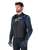 Alpinestars T-Jaws V4 Waterproof Jacket, motorjas, zwart met donkerblauw en Blithe Blue, gedragen door een model met bijpassende handschoenen en een zwarte broek, witte Alpinestars-logo's op de borst en schouders met blauwe accenten op de mouwen, sportieve textiele motorkleding