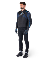 Alpinestars T-Jaws V4 Waterproof Jacket in zwart, donkerblauw en Blithe Blue, gedragen door een model met een zwarte motorbroek en motorlaarzen, voorzien van een wit Alpinestars-logo op de borst en blauwe schouderaccenten, sportieve motoruitrusting