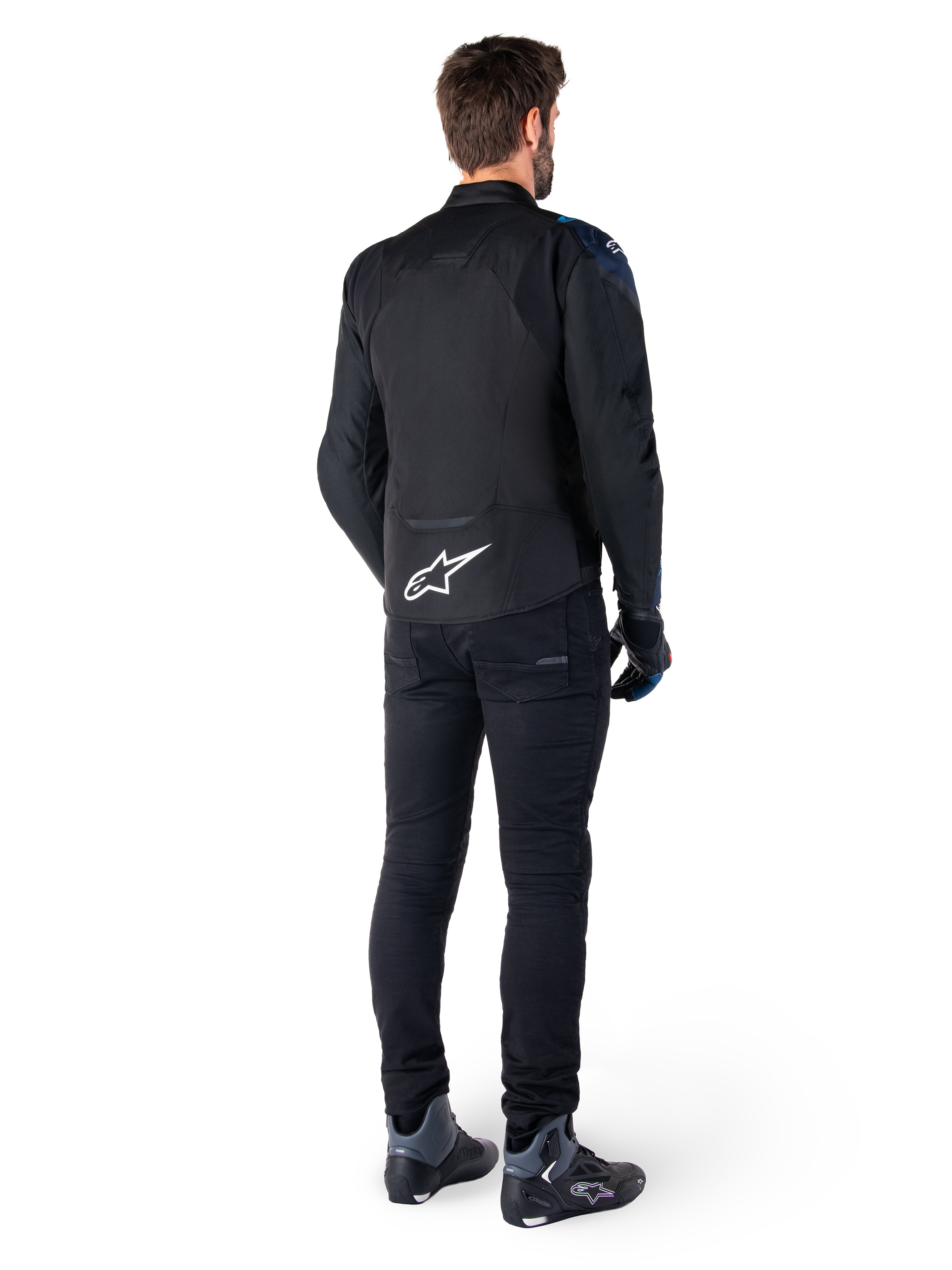 Achteraanzicht van een man die het Alpinestars T-Jaws V4 Waterproof Jacket draagt in het zwart, donkerblauw en Blithe Blue, met een opvallend wit Astar-logo op de onderrug, gecombineerd met een zwarte broek en motorschoenen.