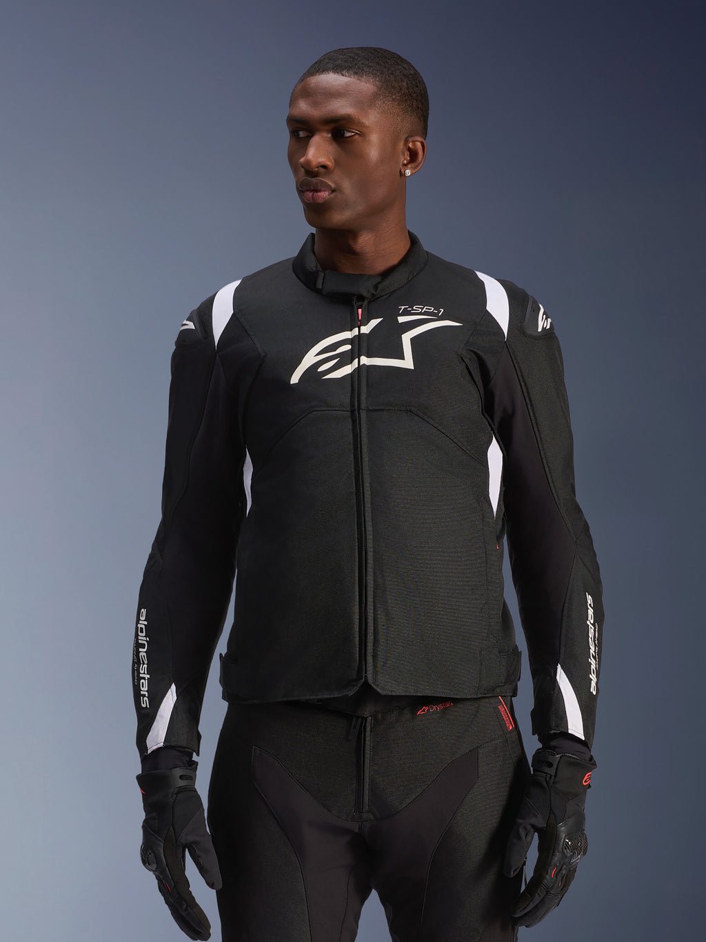 Alpinestars T-SP 1 V2 Waterproof Jacket, sportieve textiel motorjas, zwart en wit met subtiele rode details, gedragen door een mannelijk model met bijpassende handschoenen en broek, voorzien van wit Alpinestars-logo op de borst, T-SP-1-branding en beschermend silhouet voor rijden op de weg