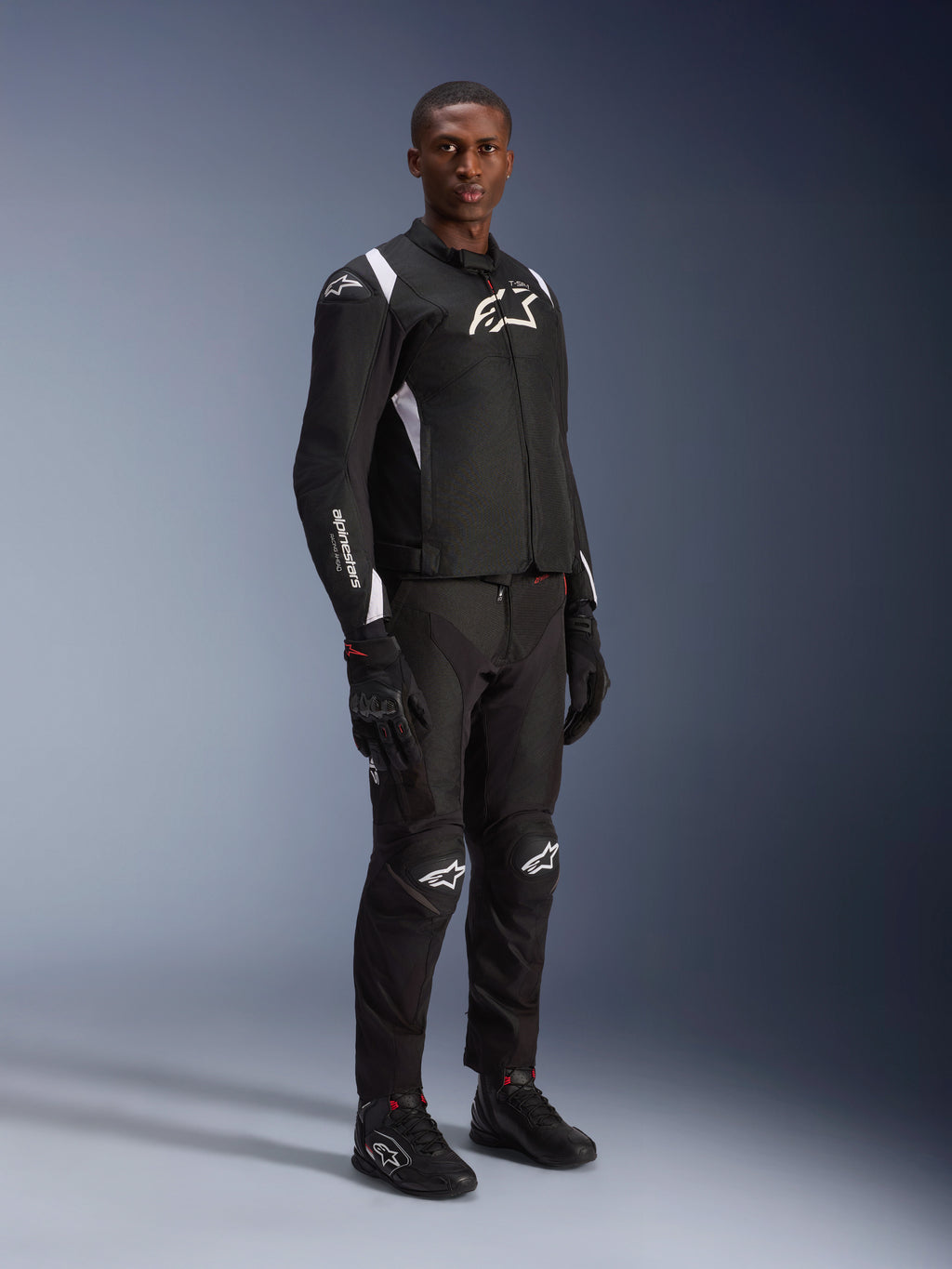 Mannelijk model draagt Alpinestars T-SP 1 V2 Waterproof Jacket in zwart en wit, gecombineerd met bijpassende zwarte motorbroek, handschoenen en laarzen. Full-body studio-opname van een sportieve textieljas met witte logo's en een 3D-getailleerd silhouet.