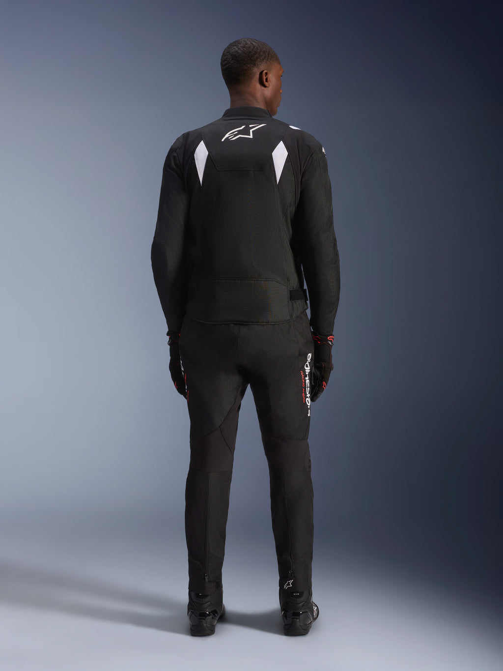 Alpinestars T-SP 1 V2 Waterproof Jacket, zwart en wit, achteraanzicht van een rijder met bijpassende zwarte motorbroek en laarzen, wit A-star-logo op de bovenrug, reflecterende schouderpanelen, sportieve textiele motorkleding