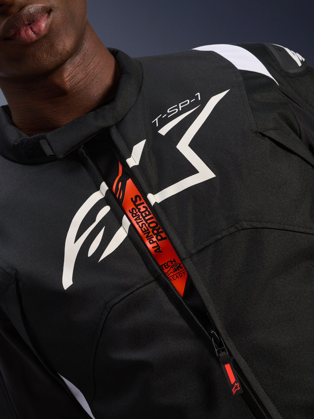 Alpinestars T-SP 1 V2 Waterdichte Jas, zwart en wit, met een groot wit logo op de borst en 'T-SP-1' tekst, oranje binnenflap met 'Alpinestars Protects' en 'Tech-Air Ready' branding, duurzame textiele motorkleding