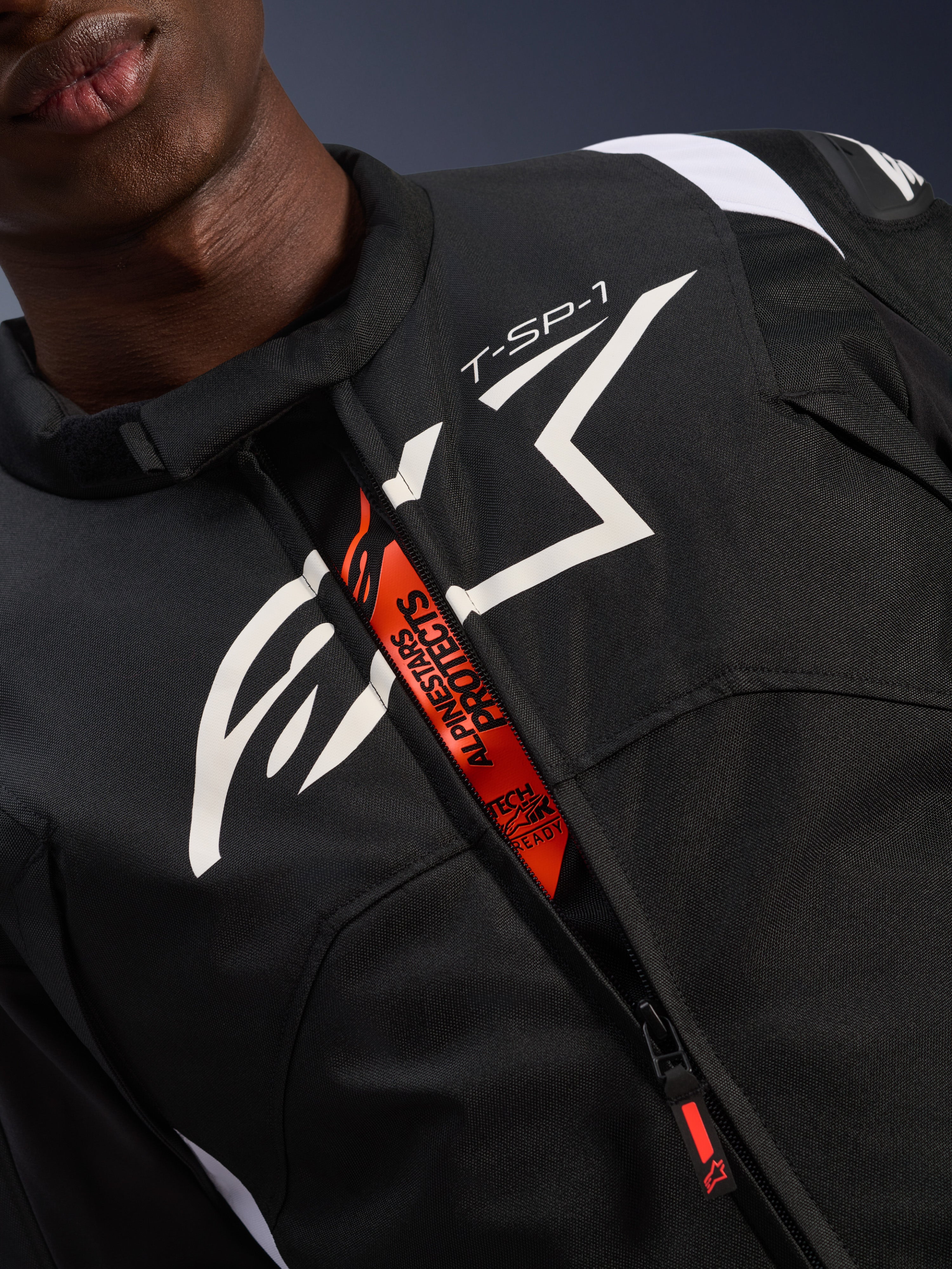 Alpinestars T-SP 1 V2 Waterdichte Jas, zwart en wit, met een groot wit logo op de borst en 'T-SP-1' tekst, oranje binnenflap met 'Alpinestars Protects' en 'Tech-Air Ready' branding, duurzame textiele motorkleding