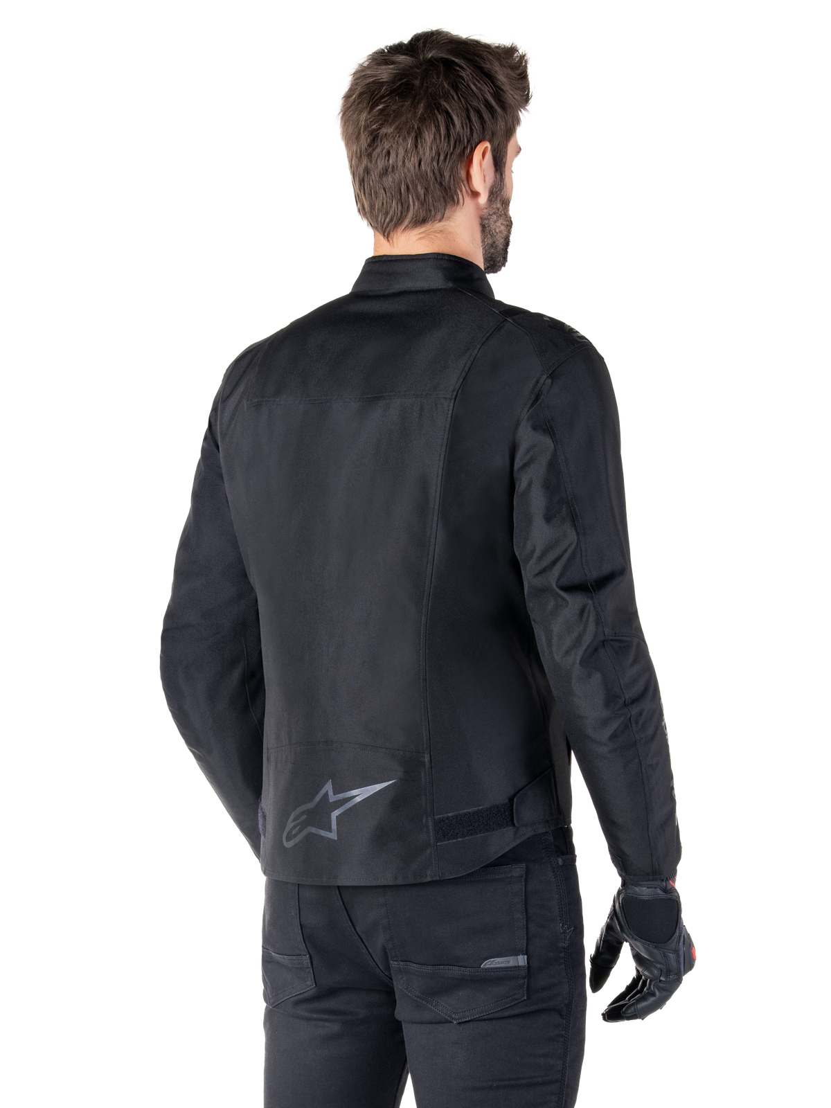 Alpinestars T-SPS V2 Waterproof Jas, zwart, achteraanzicht op een mannelijk model, met een reflecterend sterlogo op de onderrug, een verlengd rugprofiel en verstelbare taillebanden, gecombineerd met een zwarte motorbroek en handschoenen