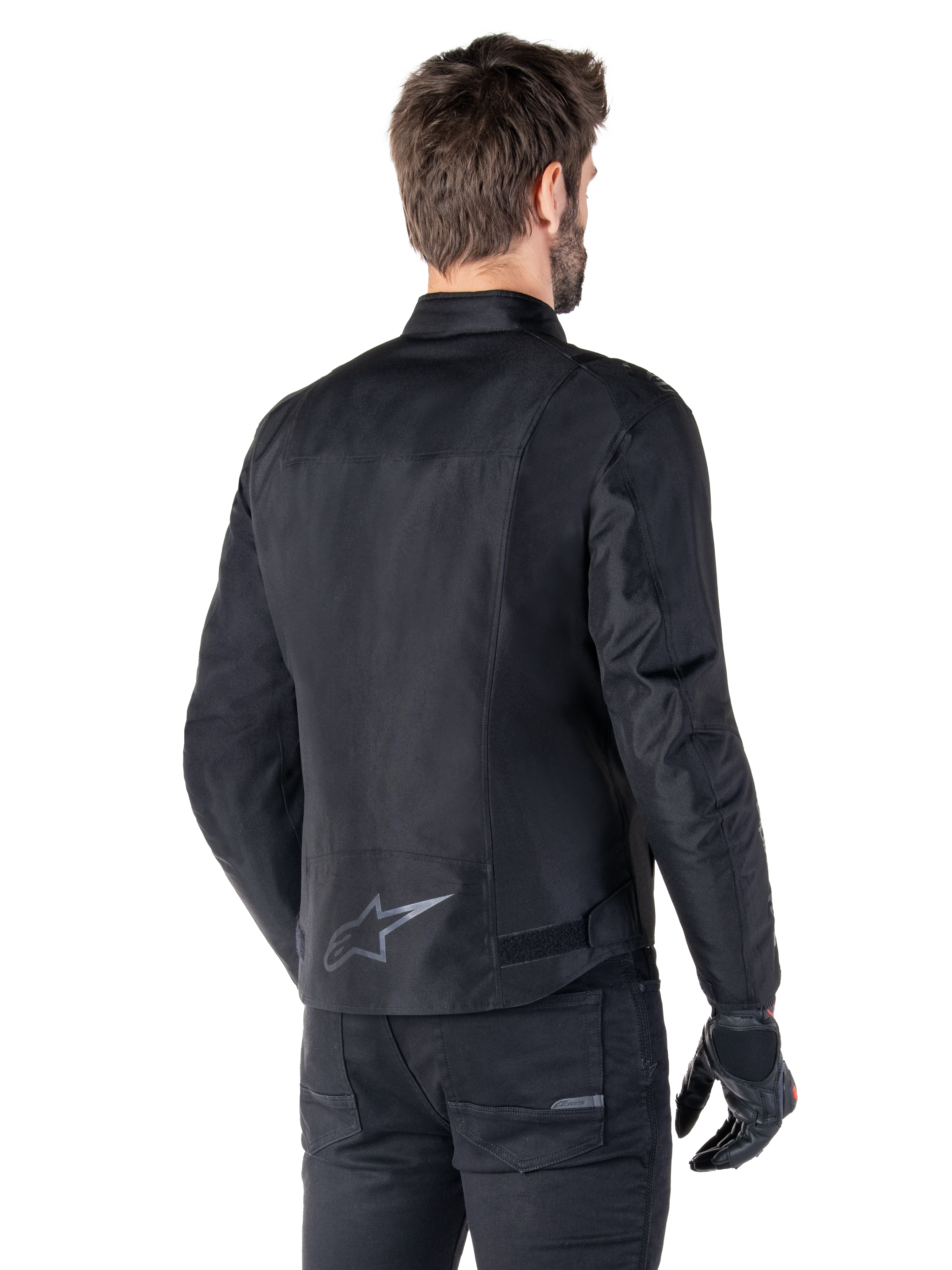 Alpinestars T-SPS V2 Waterproof Jacket, Black, rear view on a male model, featuring a reflective star logo on the lower back, extended back profile, and waist adjustment straps, paired with black riding pants and gloves