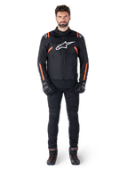 Mannelijk model draagt de Alpinestars T-SPS V2 Waterproof Jacket in Black White Red Fluo, gecombineerd met een zwarte motorbroek en handschoenen, voorzien van een wit Alpinestars-logo op de borst en rode accenten op de mouwen.