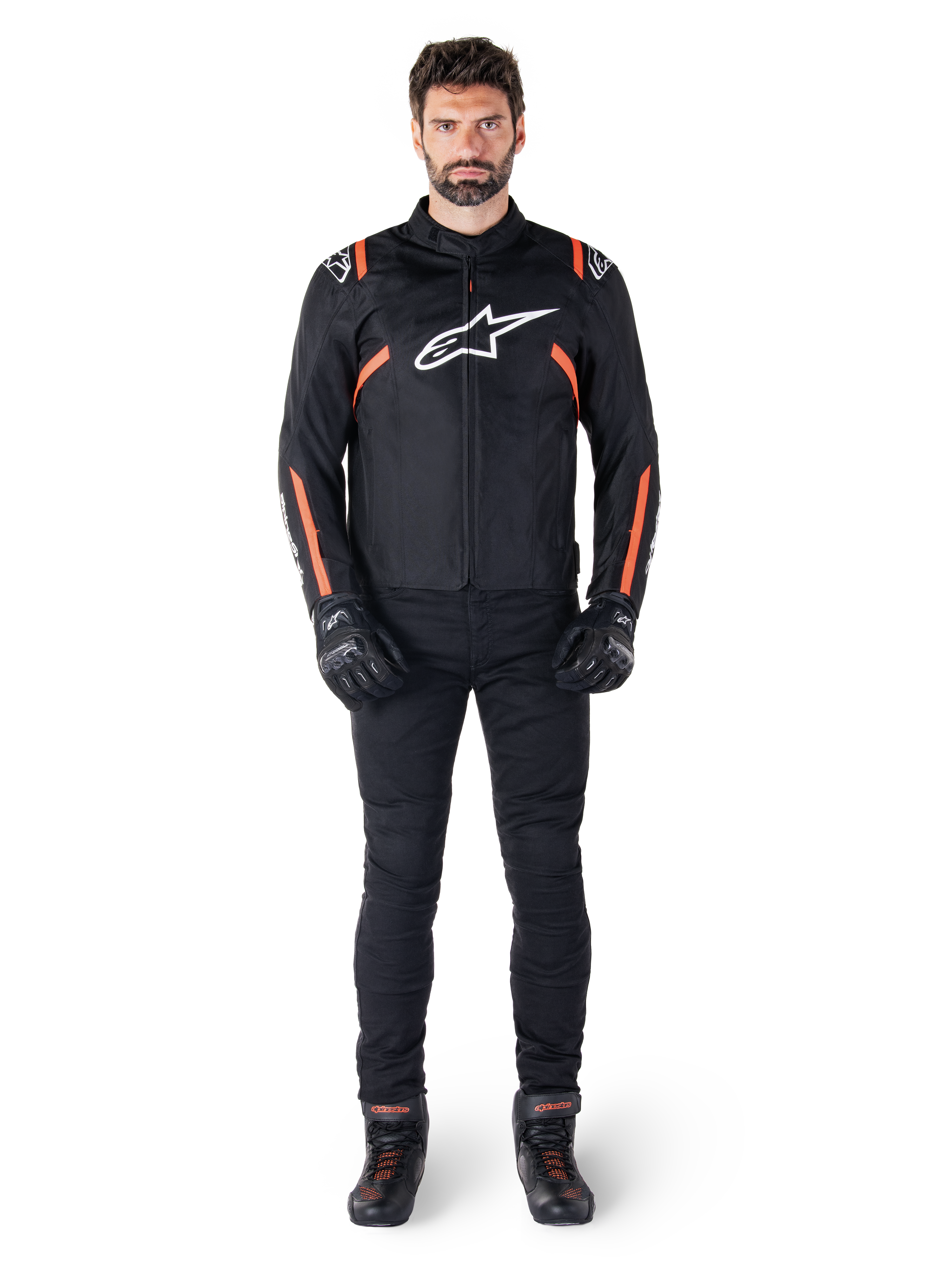 Mannelijk model draagt de Alpinestars T-SPS V2 Waterproof Jacket in Black White Red Fluo, gecombineerd met een zwarte motorbroek en handschoenen, voorzien van een wit Alpinestars-logo op de borst en rode accenten op de mouwen.