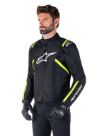 Mannelijk model draagt de Alpinestars T-SPS V2 Waterproof Jas, zwart met witte en fluorgele accenten, voorzien van een wit Astars-logo op de borst en fluorescerend gele strepen op de schouders en mouwen, gecombineerd met een zwarte broek en motorhandschoenen, sportief gesneden textiel motorjas.
