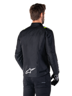 Alpinestars T-SPS V2 Waterproof Jacket, zwart wit fluogeel, achteraanzicht van een mannelijk model dat de textiele motorjas draagt, met een wit logo op de onderrug en fluogele accenten op de schouders, gecombineerd met een zwarte broek en handschoenen.