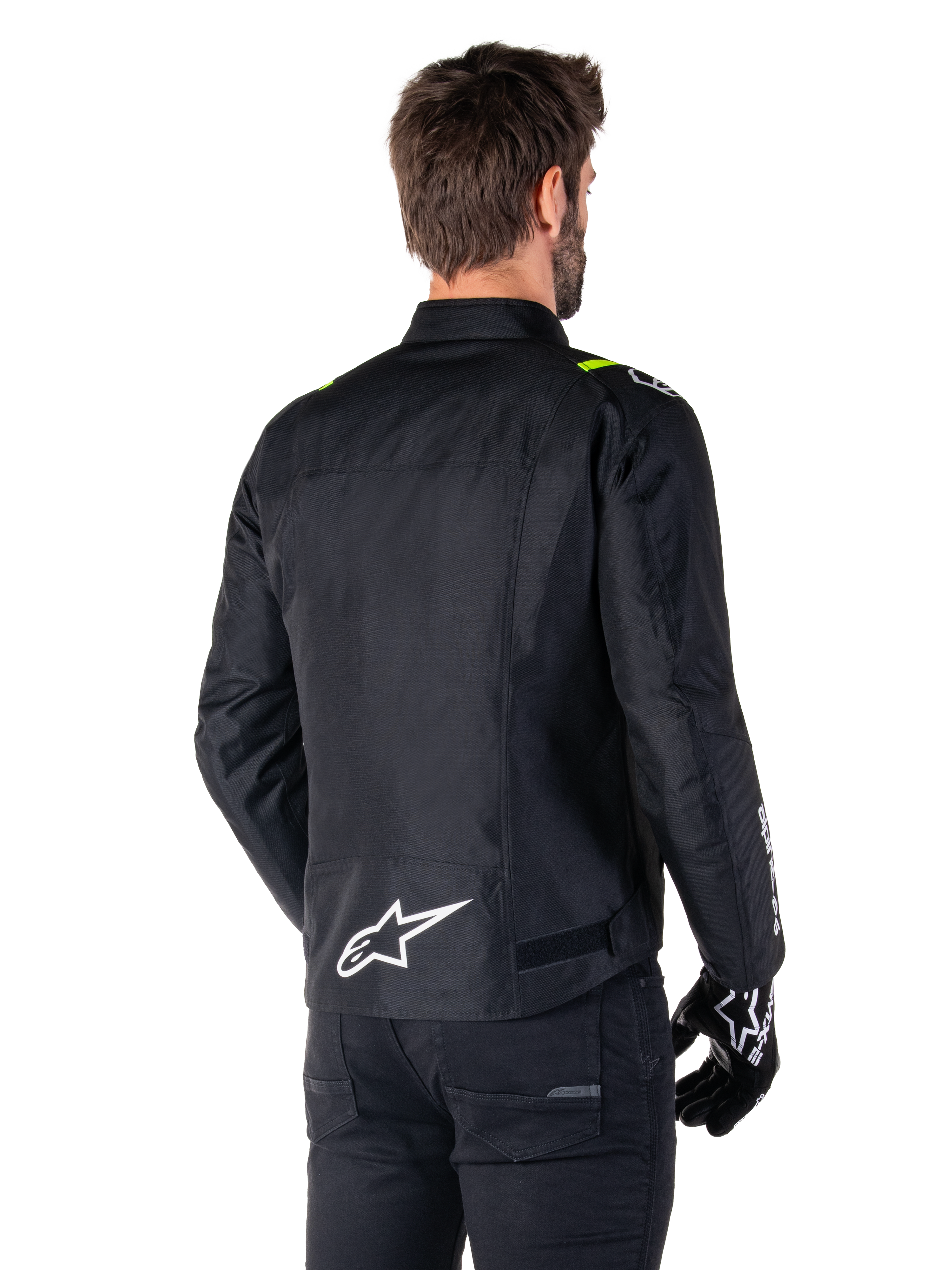 Alpinestars T-SPS V2 Waterproof Jacket, zwart wit fluogeel, achteraanzicht van een mannelijk model dat de textiele motorjas draagt, met een wit logo op de onderrug en fluogele accenten op de schouders, gecombineerd met een zwarte broek en handschoenen.
