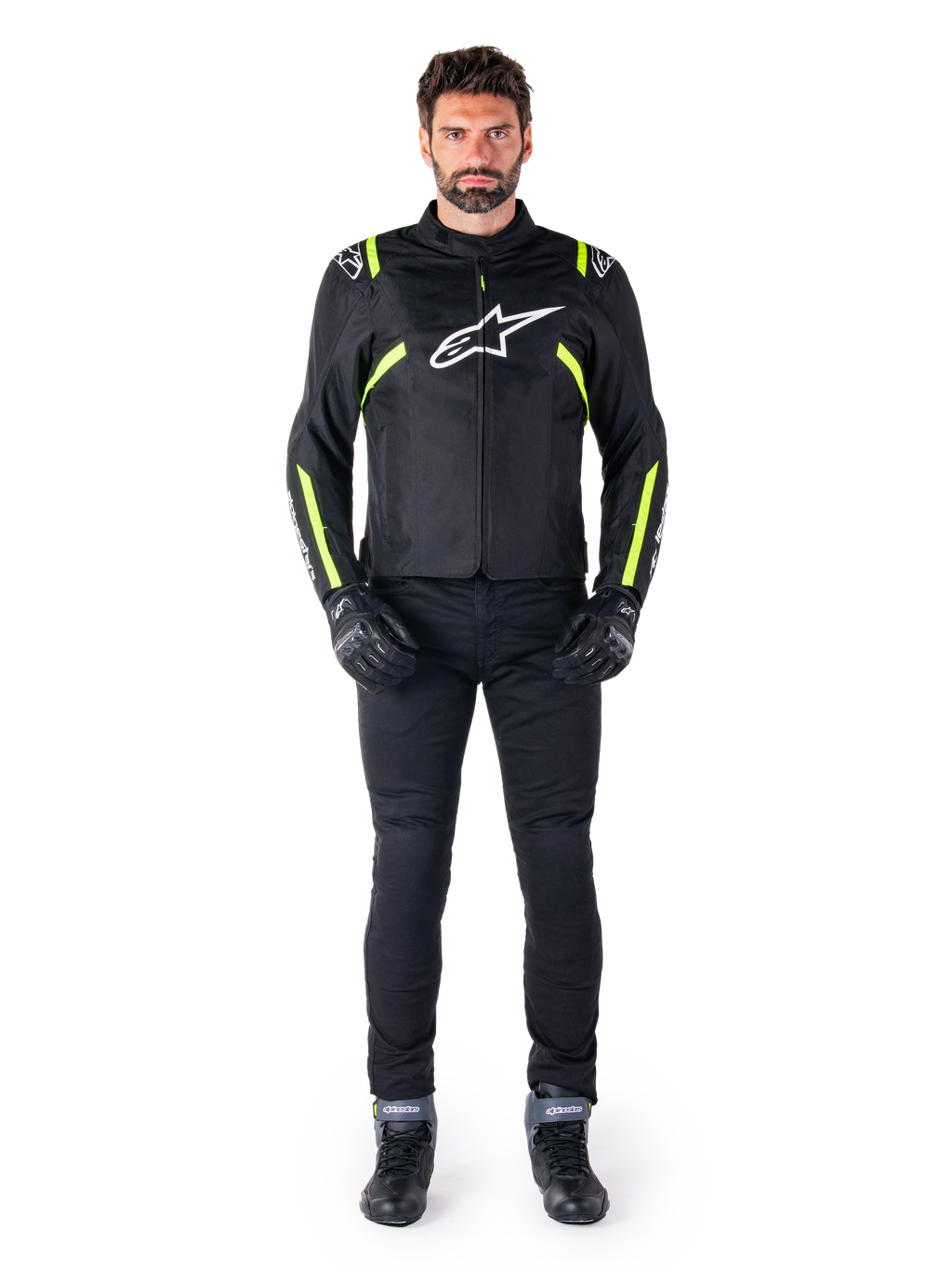 Alpinestars T-SPS V2 Waterproof jas, zwart, wit en geel fluo, gedragen door een model met zwarte motorbroek en handschoenen, wit Alpinestars logo op de borst en fluorescerende accenten op de schouders en mouwen, sportieve motoruitrusting