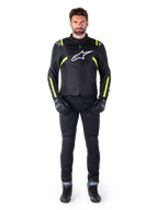 Alpinestars T-SPS V2 Waterproof jas, zwart, wit en geel fluo, gedragen door een model met zwarte motorbroek en handschoenen, wit Alpinestars logo op de borst en fluorescerende accenten op de schouders en mouwen, sportieve motoruitrusting