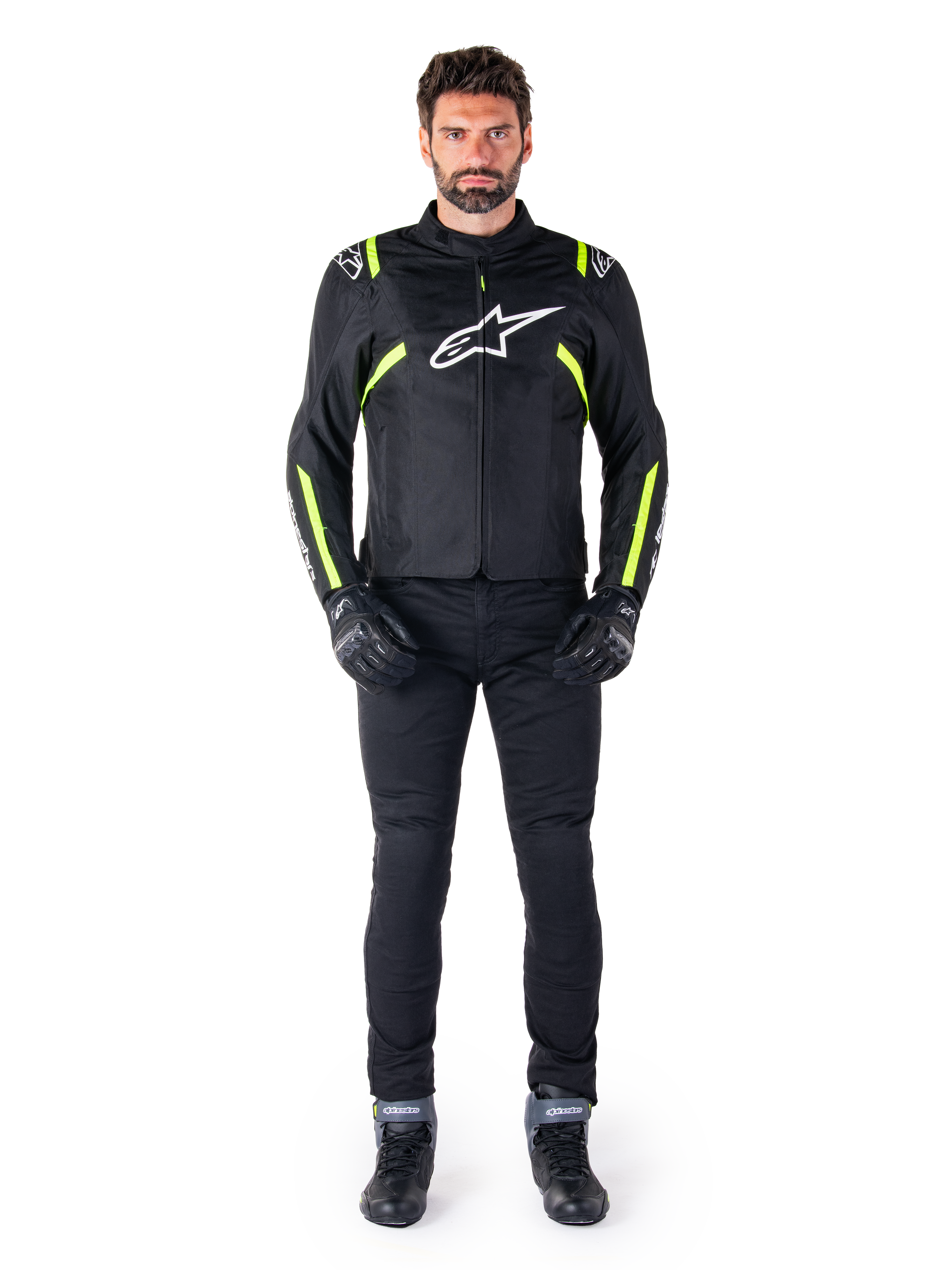 Alpinestars T-SPS V2 Waterproof jas, zwart, wit en geel fluo, gedragen door een model met zwarte motorbroek en handschoenen, wit Alpinestars logo op de borst en fluorescerende accenten op de schouders en mouwen, sportieve motoruitrusting