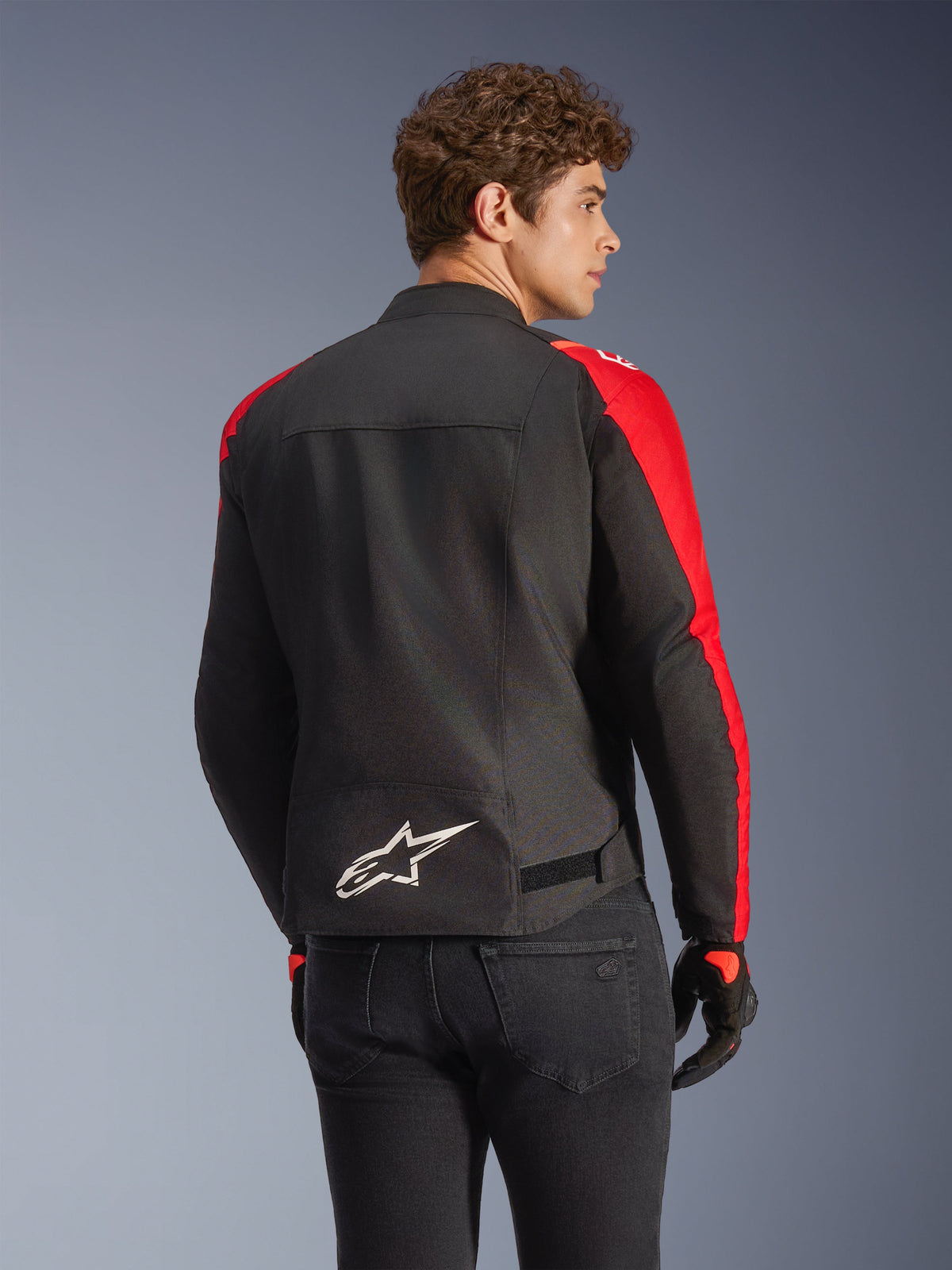 Alpinestars T-SPS V2 Waterproof Jas in zwart en rood, achteraanzicht op een model, wit Alpinestars-logo op de onderrug, rode mouwpanelen, sportief silhouet, gedragen met zwarte handschoenen en spijkerbroek.
