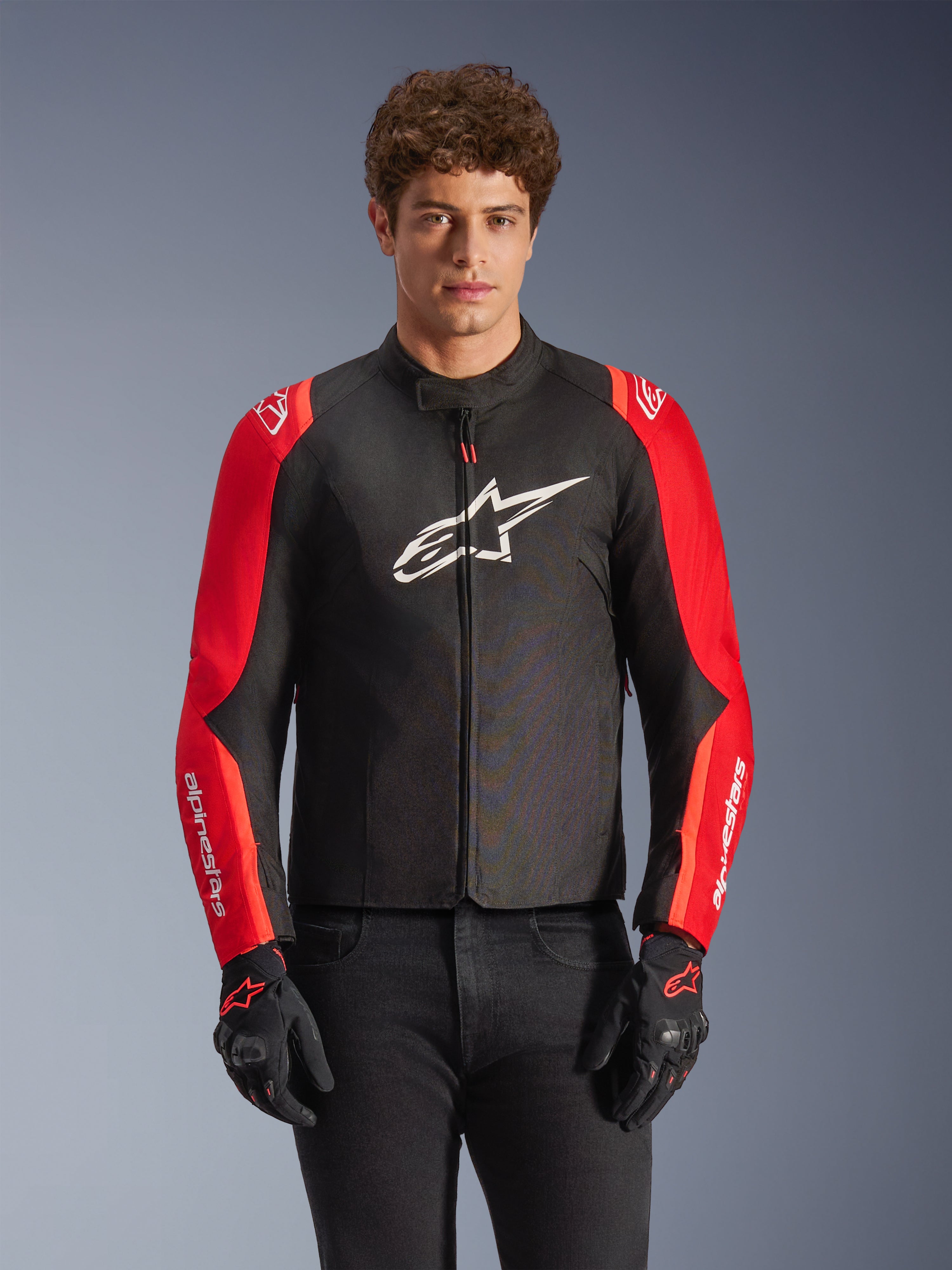 Alpinestars T-SPS V2 Waterproof Jas, textiele motorjas, zwart en rood, gedragen door een mannelijk model met bijpassende zwarte handschoenen, met een groot wit logo op de borst en witte merkdetails op de rode mouwen, sportief silhouet voor rijden in alle weersomstandigheden