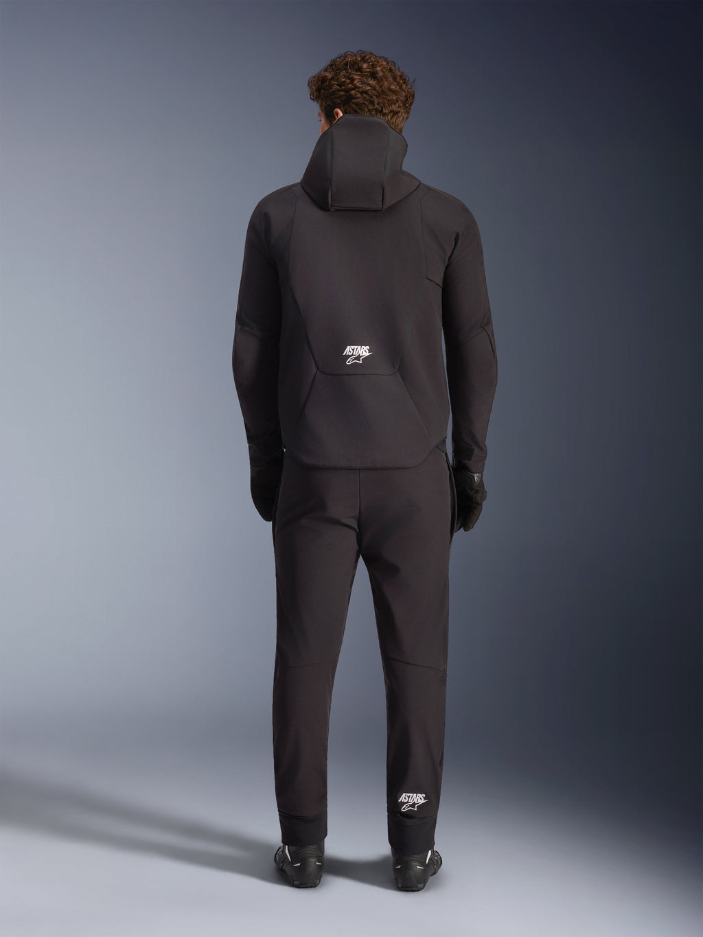 Alpinestars Aeroshell Waterproof Jacket, Black, seen from the back on a model wearing matching black riding pants and gloves, hooded sports softshell design with white ASTARS logos, sleek and modern motorcycle apparel