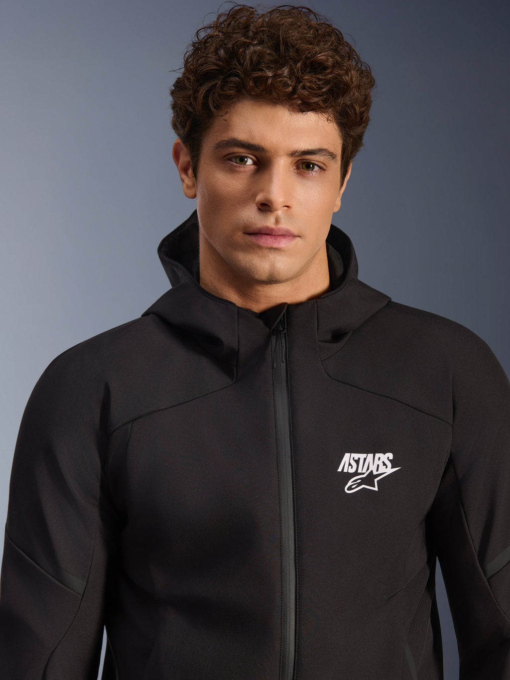 Male model wearing Alpinestars Aeroshell Waterproof Jacket, black textile softshell hoodie, white ASTARS logo on chest, waterproof front zipper, modern studio portrait for motorcycle road gear.