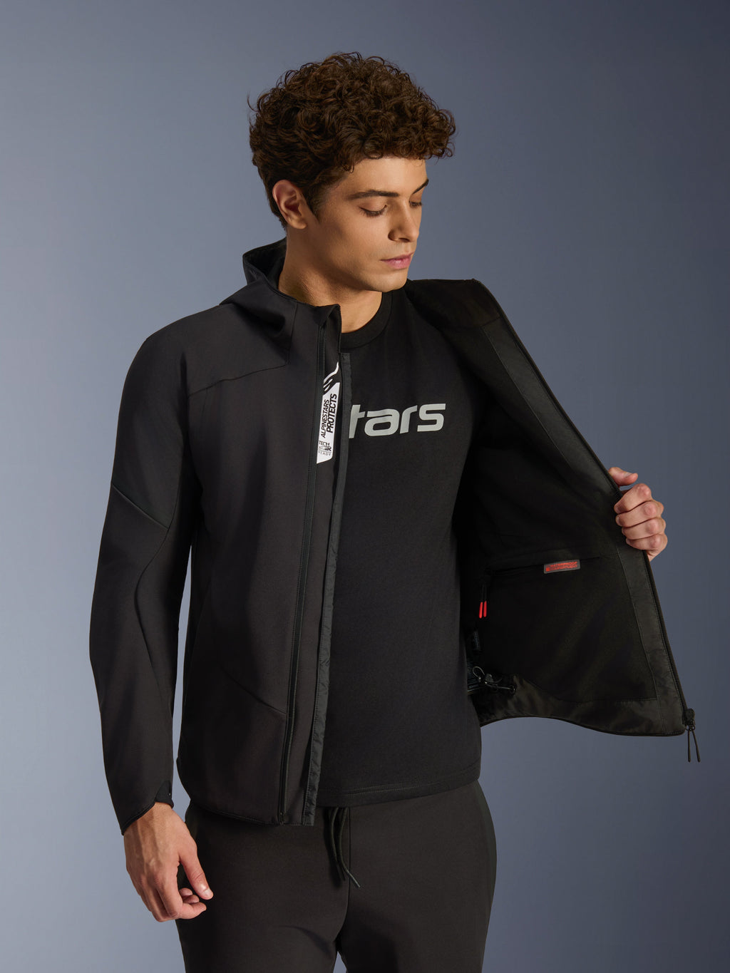 Alpinestars Aeroshell Waterproof Jacket, Black softshell motorcycle jacket, unzipped showing interior pocket and lining, worn by a male model with a black logo tee, hooded design for casual urban riding and street style.
