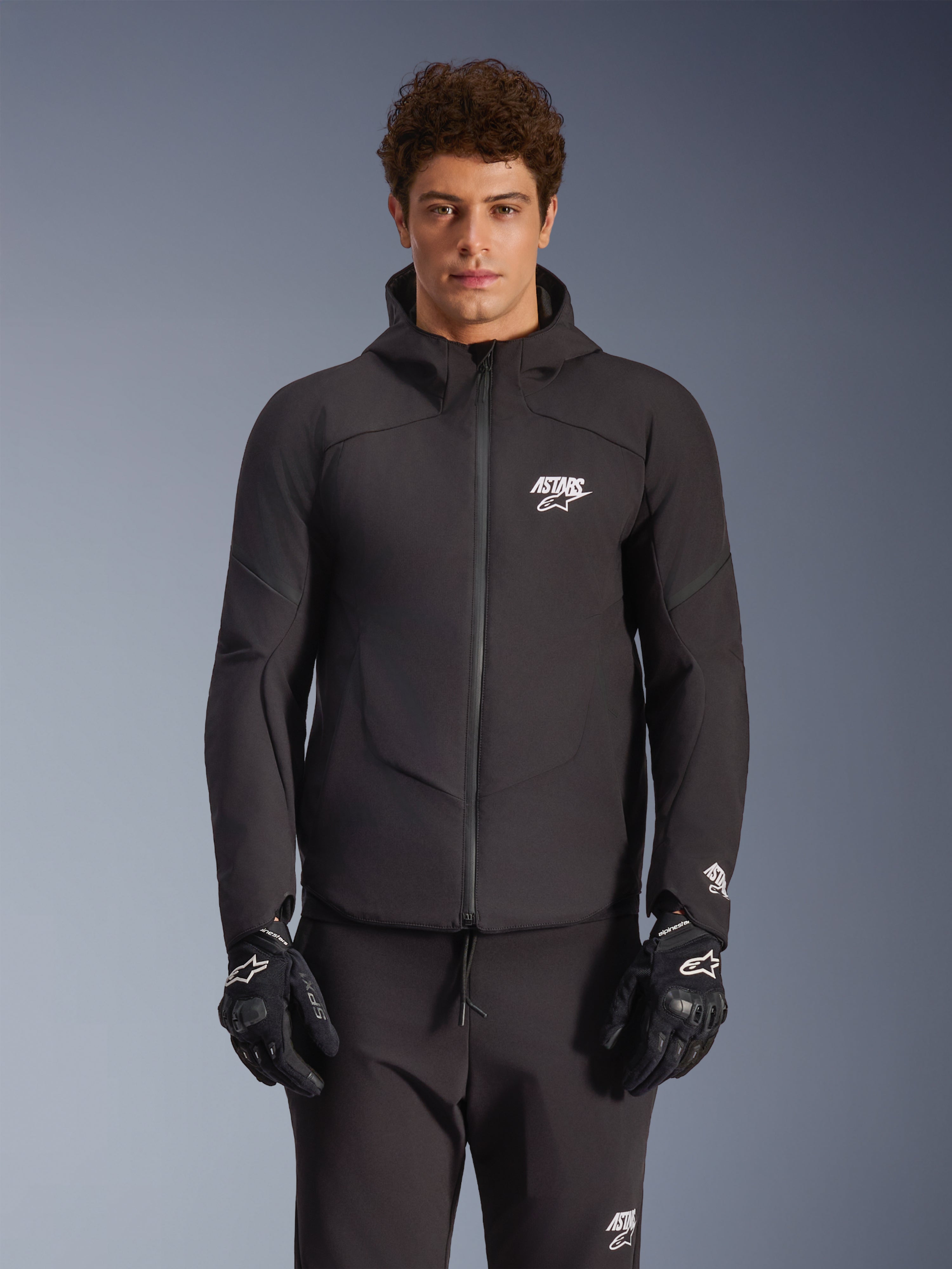 Alpinestars Aeroshell Waterproof Jacket, zwarte urban motorjas, ontwerp met capuchon en volledige ritssluiting, witte logo's op de borst en mouwen, gedragen met bijpassende zwarte broek en handschoenen