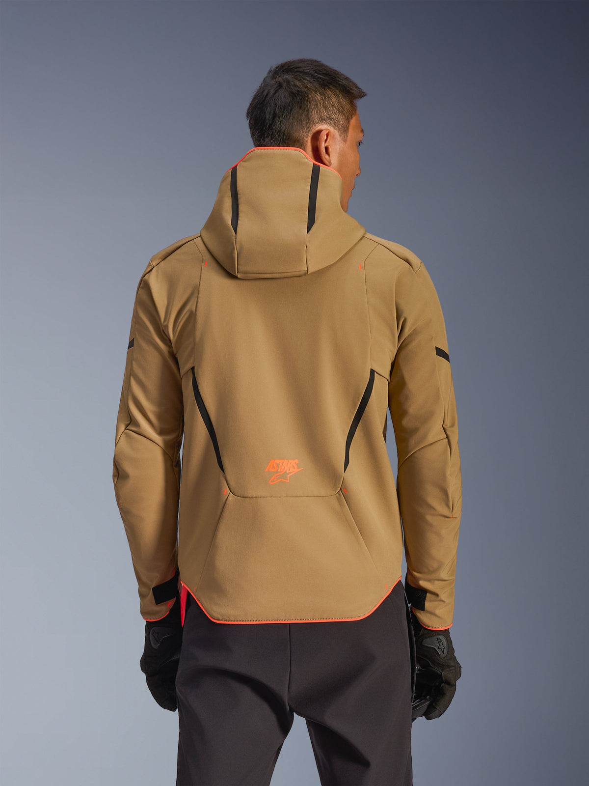 Alpinestars Aeroshell Waterproof Jacket, Dark Khaki en Orange Fluo, achteraanzicht met hoodie-ontwerp, oranje bies op de capuchon en zoom, oranje logo op de onderrug, gedragen met zwarte handschoenen, crossover sportieve softshell motoruitrusting.