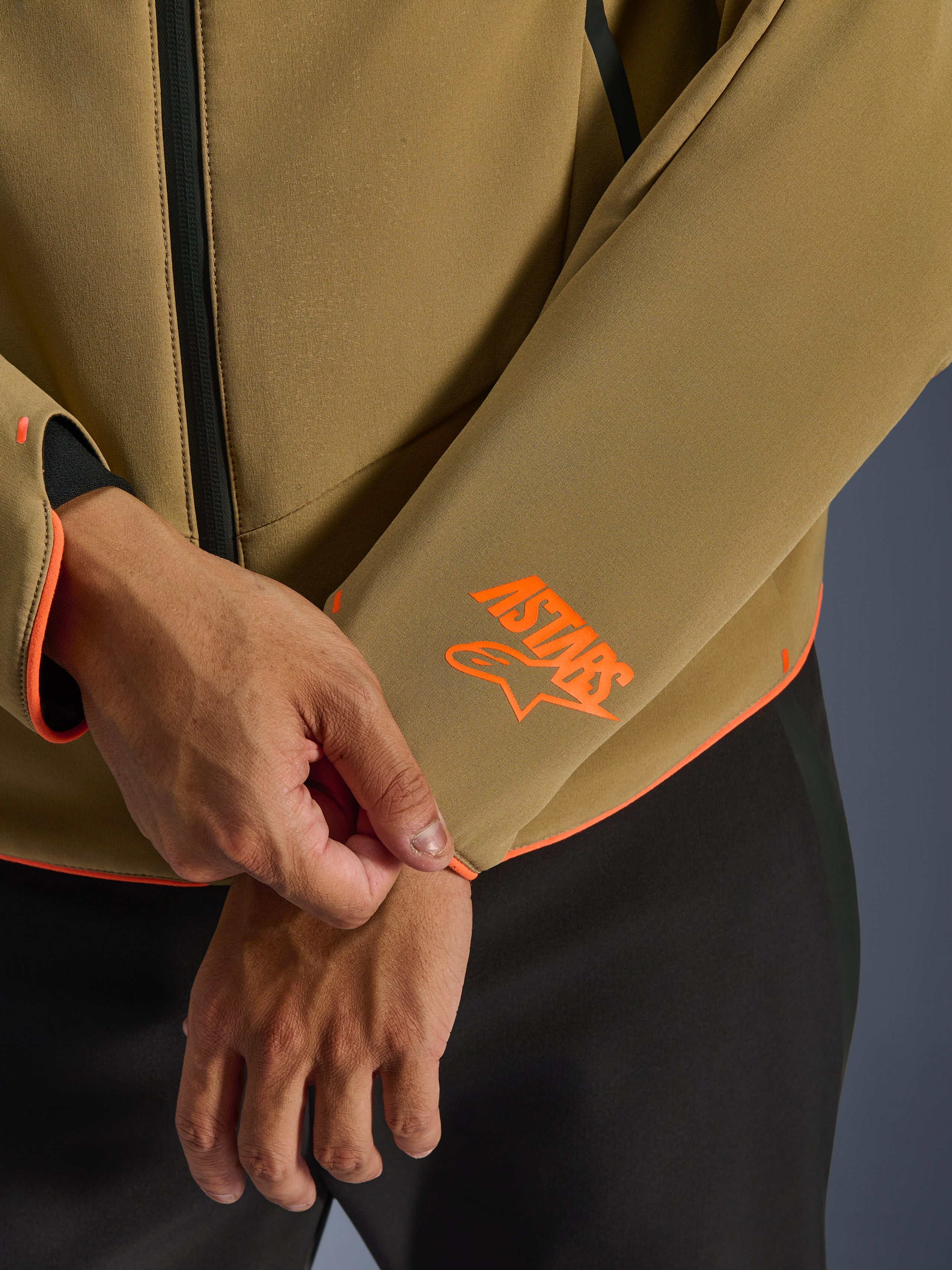 Alpinestars Aeroshell Waterproof Jacket, donker kaki met fluoriserend oranje accenten, close-up van de mouwboord en onderste zoom met het feloranje Astars-logo, neon oranje piping en detail van de waterdichte rits, sportieve softshell-constructie.
