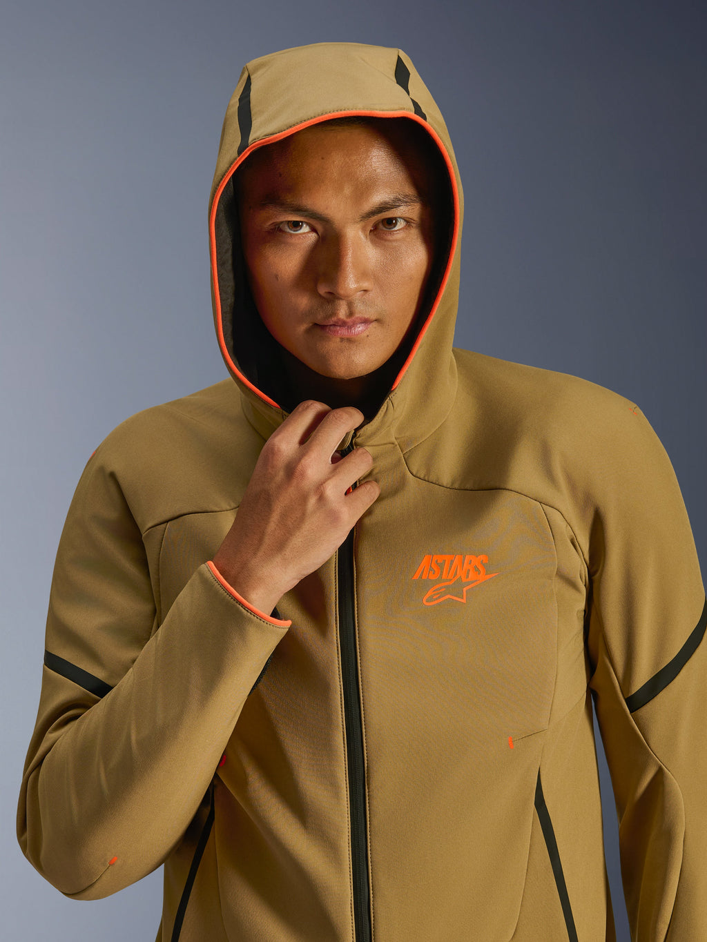 Alpinestars Aeroshell waterdichte jas, Dark Khaki Black Orange Fluo, model draagt de jas met de capuchon omhoog, voorzien van opvallende oranje fluo details en een oranje ASTARS-logo, strakke zwarte accenten, moderne sportieve waterdichte softshell
