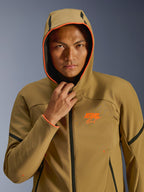 Alpinestars Aeroshell waterdichte jas, Dark Khaki Black Orange Fluo, model draagt de jas met de capuchon omhoog, voorzien van opvallende oranje fluo details en een oranje ASTARS-logo, strakke zwarte accenten, moderne sportieve waterdichte softshell