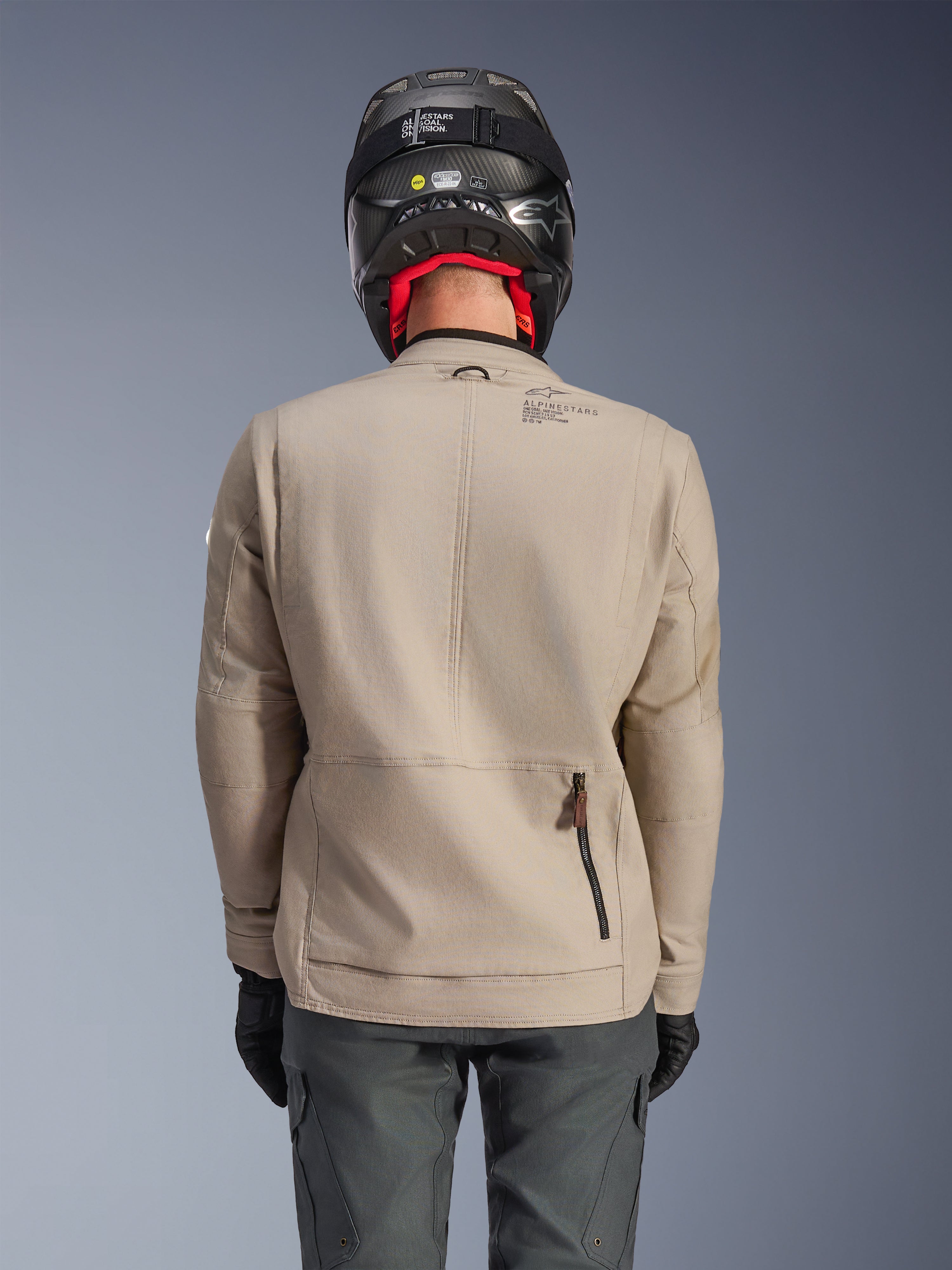 Alpinestars Borrego Drystar® Jas, adventure motorjas, warm grijs, achteraanzicht met rits van het achtercompartiment en Alpinestars-branding, gedragen met een zwarte helm en antracietkleurige motorbroek