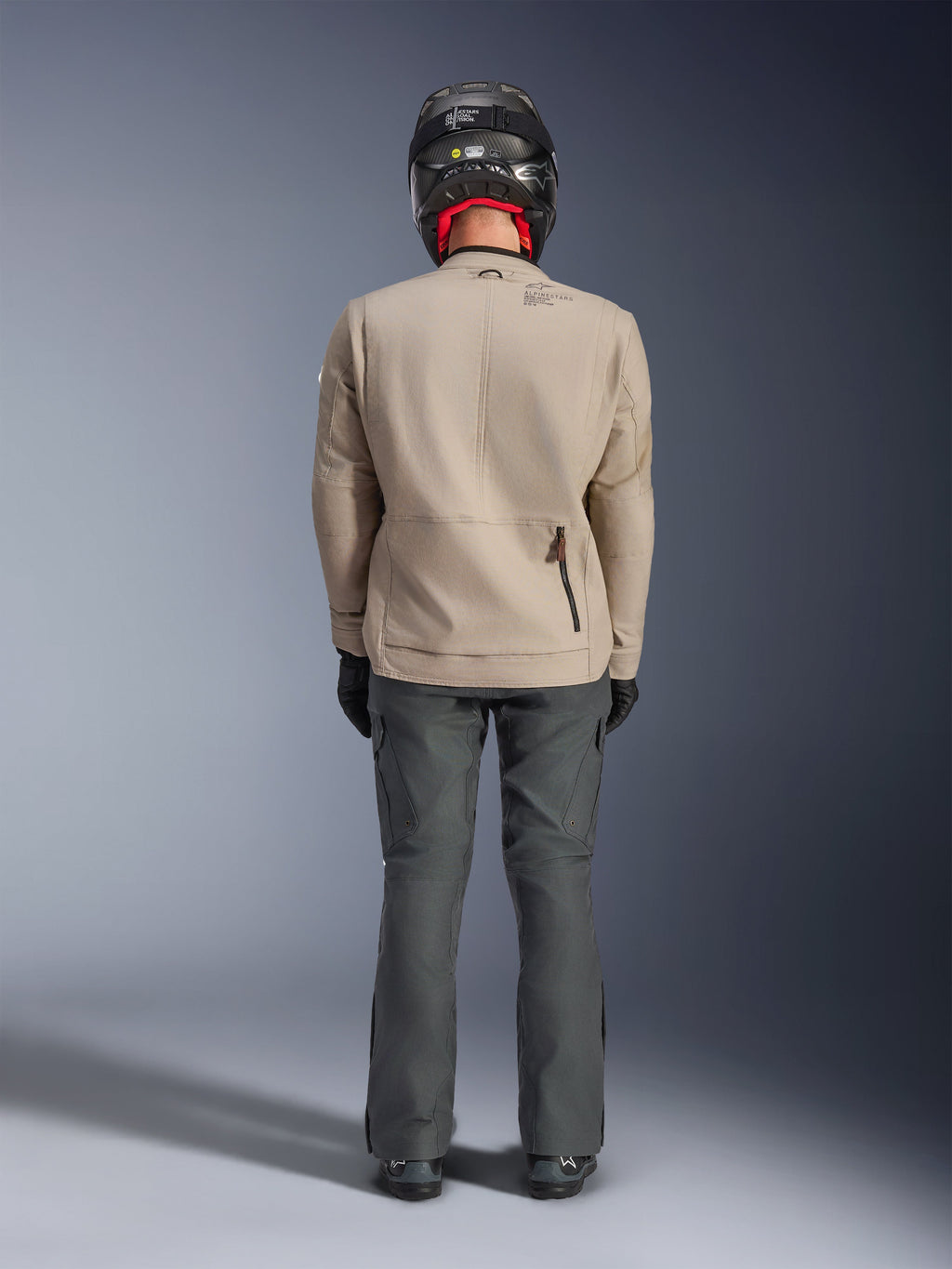 Alpinestars Borrego Drystar® Jas, Warm Gray avontuurlijke motorjas, achteraanzicht met gewaxt canvas uitstraling, schouderuitbreidingspanelen en ritszak op de onderrug, gedragen met donkergrijze motorbroek en zwarte helm.
