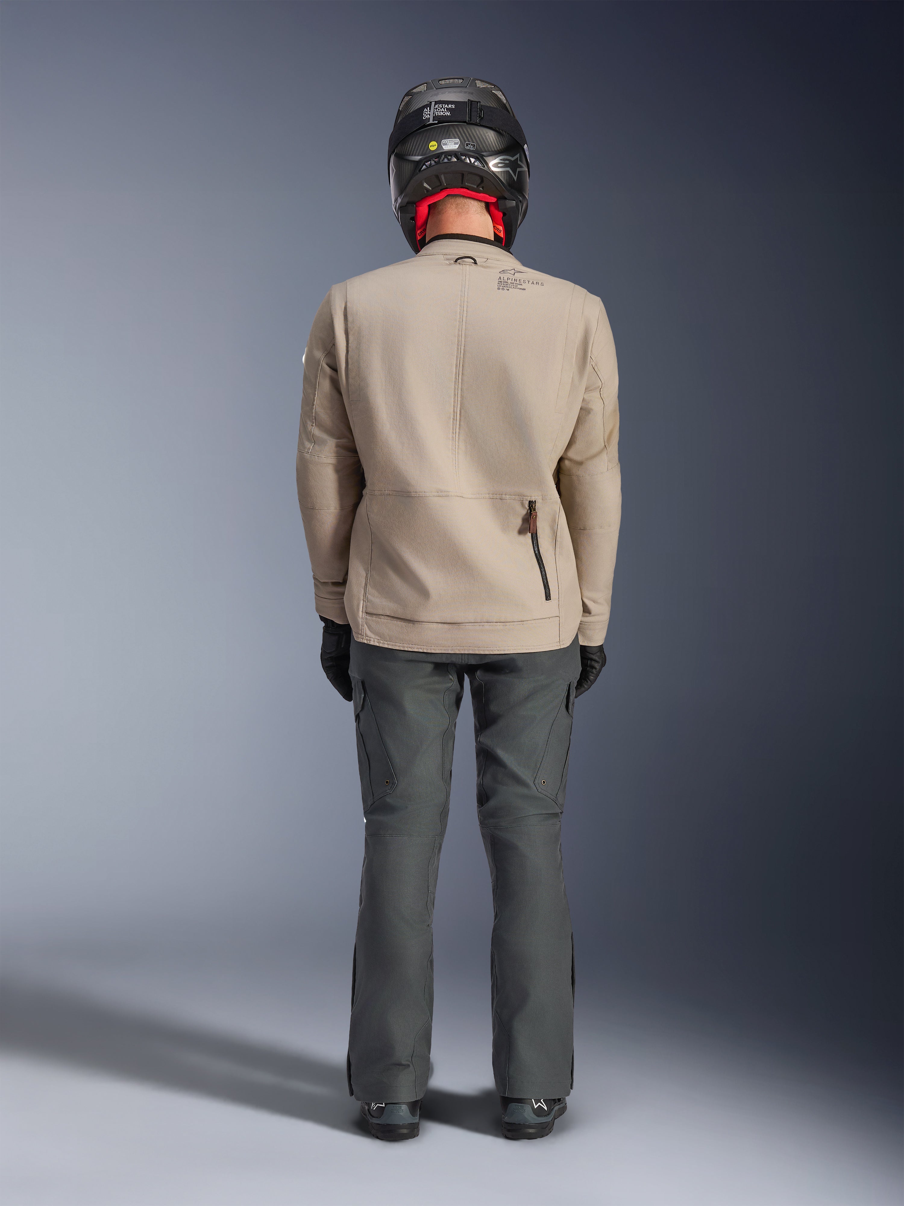 Alpinestars Borrego Drystar® Jas, Warm Gray avontuurlijke motorjas, achteraanzicht met gewaxt canvas uitstraling, schouderuitbreidingspanelen en ritszak op de onderrug, gedragen met donkergrijze motorbroek en zwarte helm.