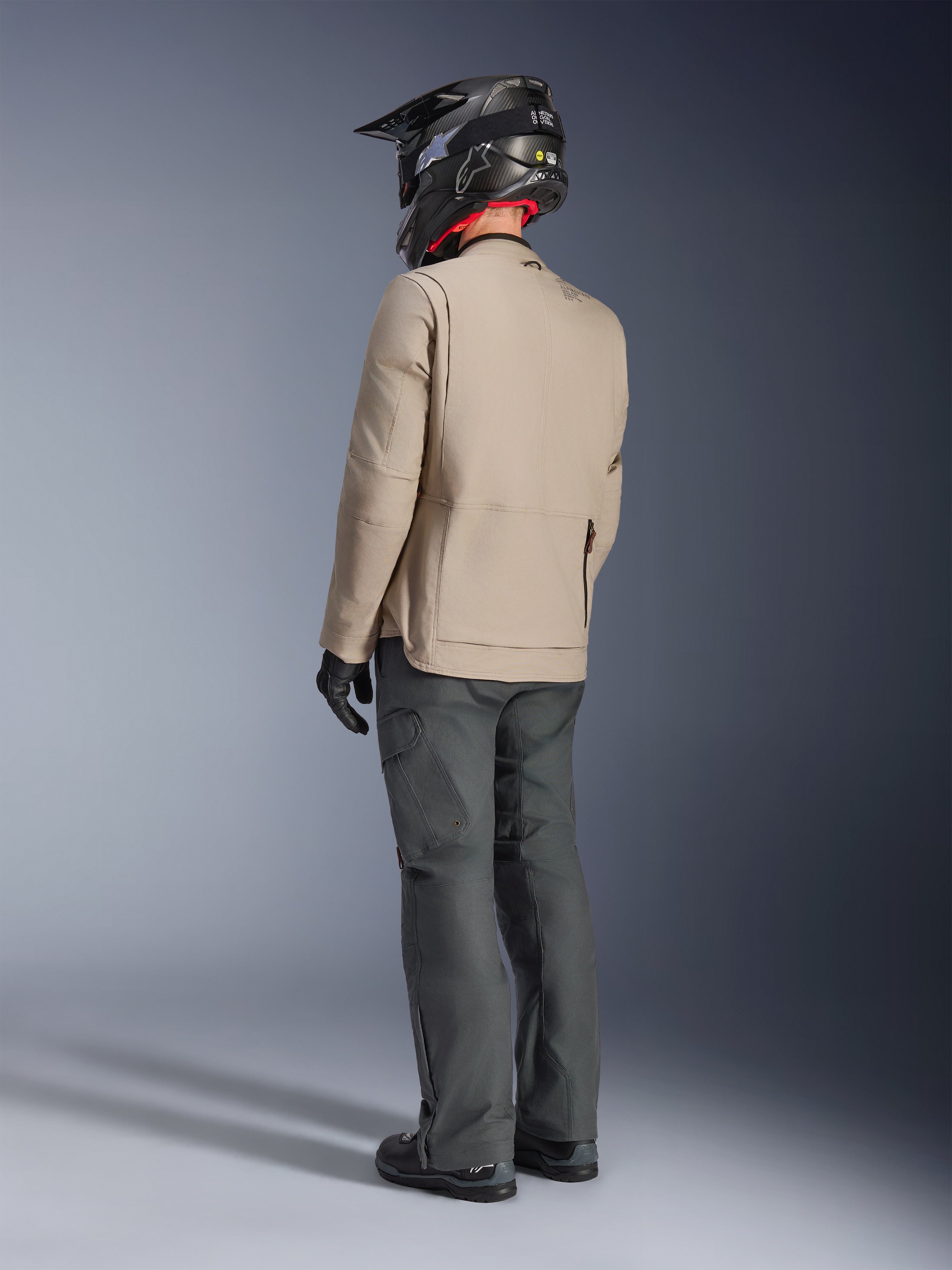 Alpinestars Borrego Drystar® Jas, Warm Gray adventure motorjas, achteraanzicht van motorrijder met donkergrijze broek en zwarte helm, voorzien van een robuuste gewaxt canvas look en functioneel ontwerp.