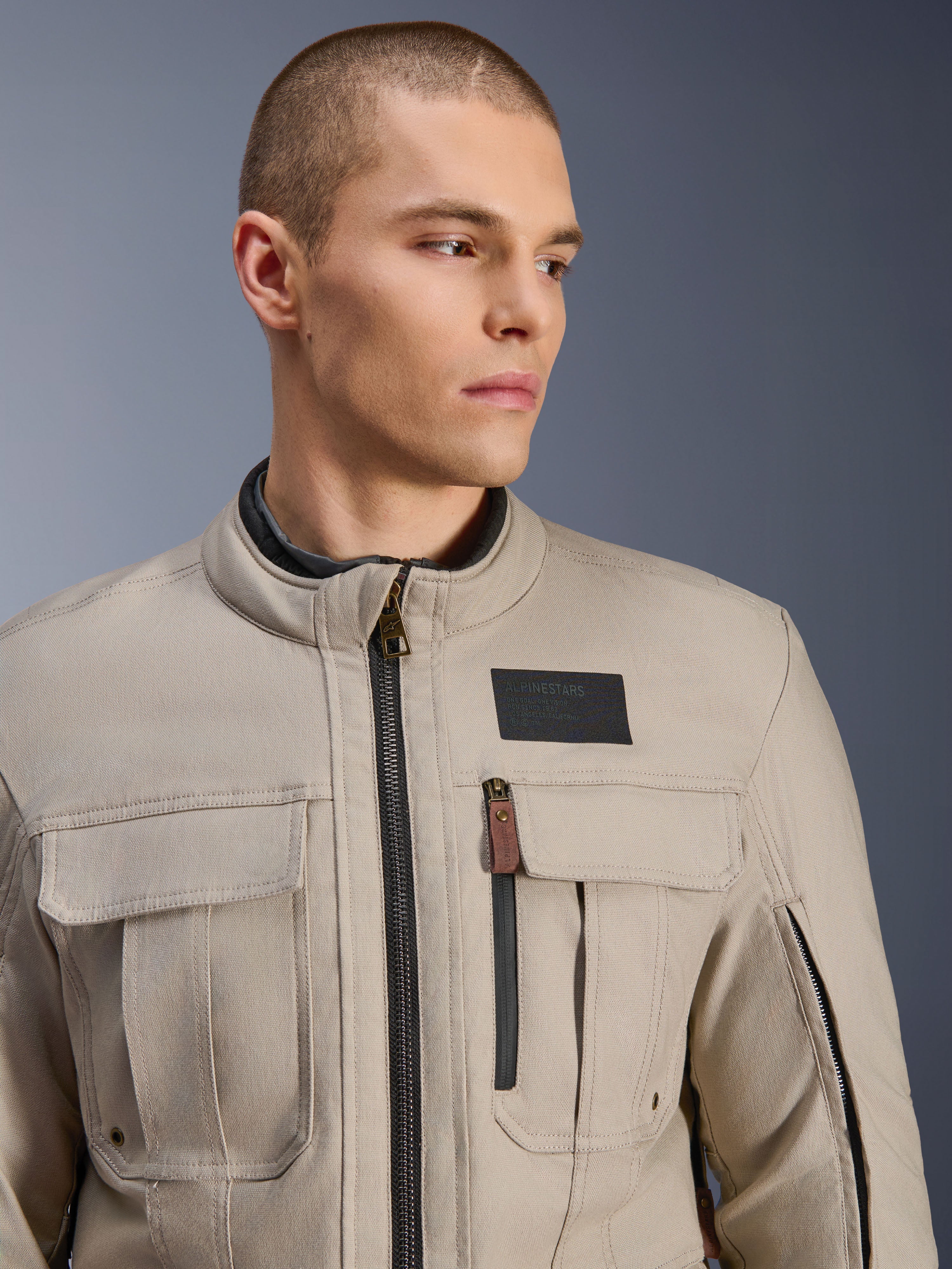 Mannelijk model draagt de Alpinestars Borrego Drystar® Jas in Warm Gray, adventure motorjas met een klassieke gewaxt canvas uitstraling, functionele cargo-borstzakken en een zwarte Alpinestars logo-patch.