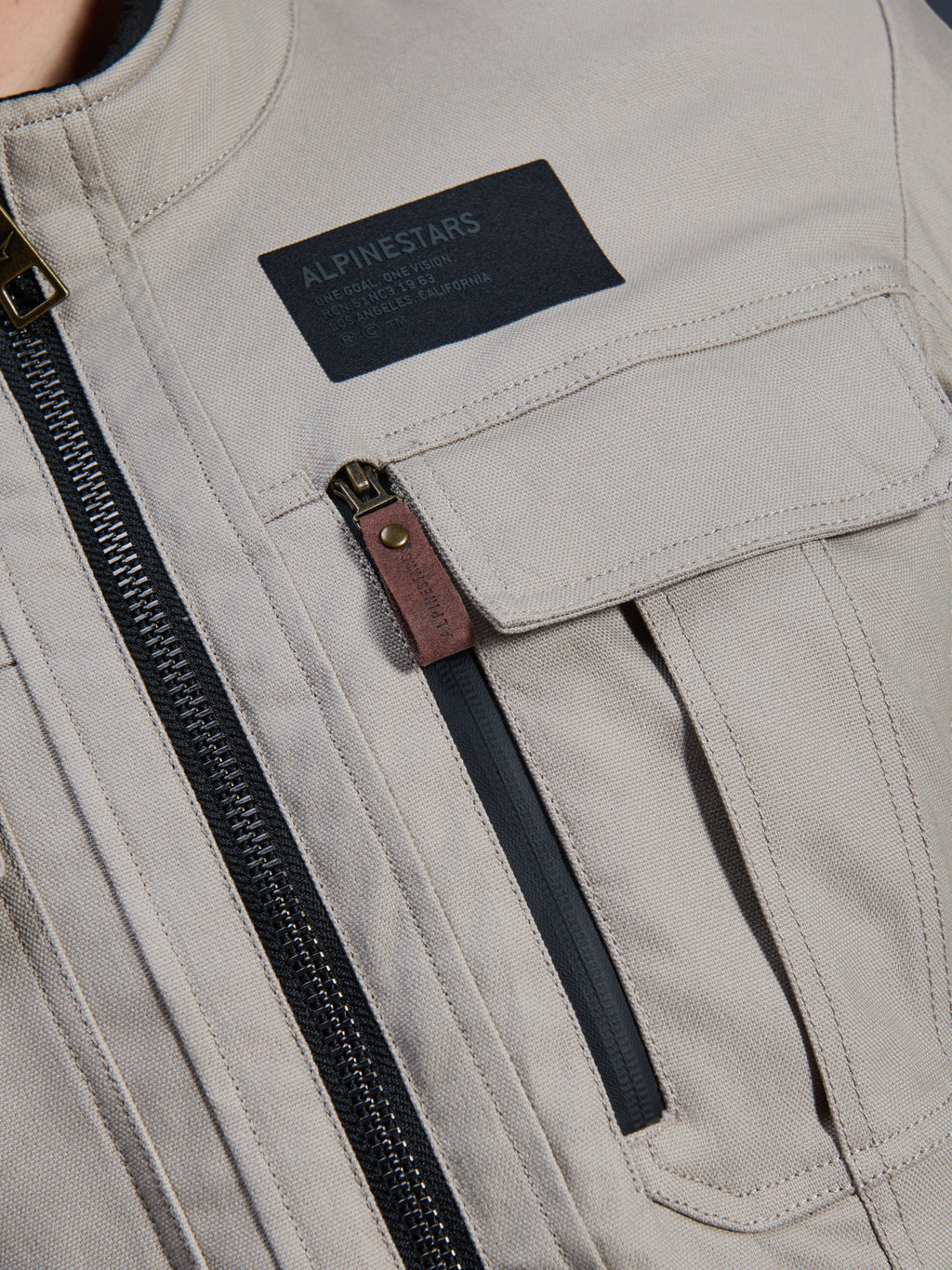 Alpinestars Borrego Drystar® Jas, Warm Gray, detailopname van de borststreek met een zwarte Alpinestars merkpatch, borstzak met klep en een verticale rits met een bruin lederen treklipje, duurzame grijze textiel canvas constructie.