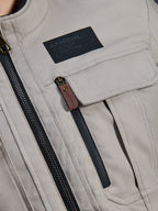 Alpinestars Borrego Drystar® Jas, Warm Gray, detailopname van de borststreek met een zwarte Alpinestars merkpatch, borstzak met klep en een verticale rits met een bruin lederen treklipje, duurzame grijze textiel canvas constructie.