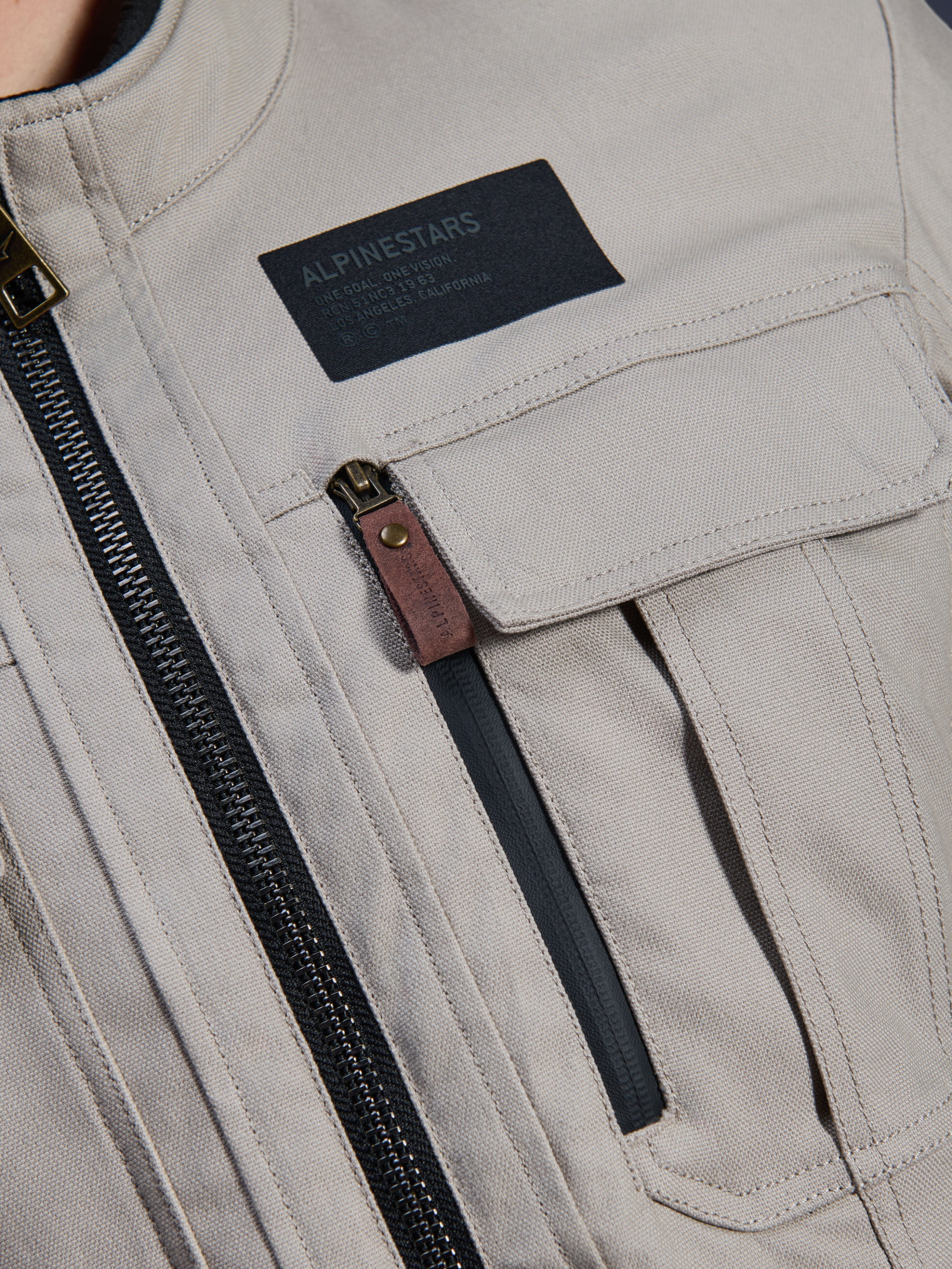 Alpinestars Borrego Drystar® Jas, Warm Gray, detailopname van de borststreek met een zwarte Alpinestars merkpatch, borstzak met klep en een verticale rits met een bruin lederen treklipje, duurzame grijze textiel canvas constructie.