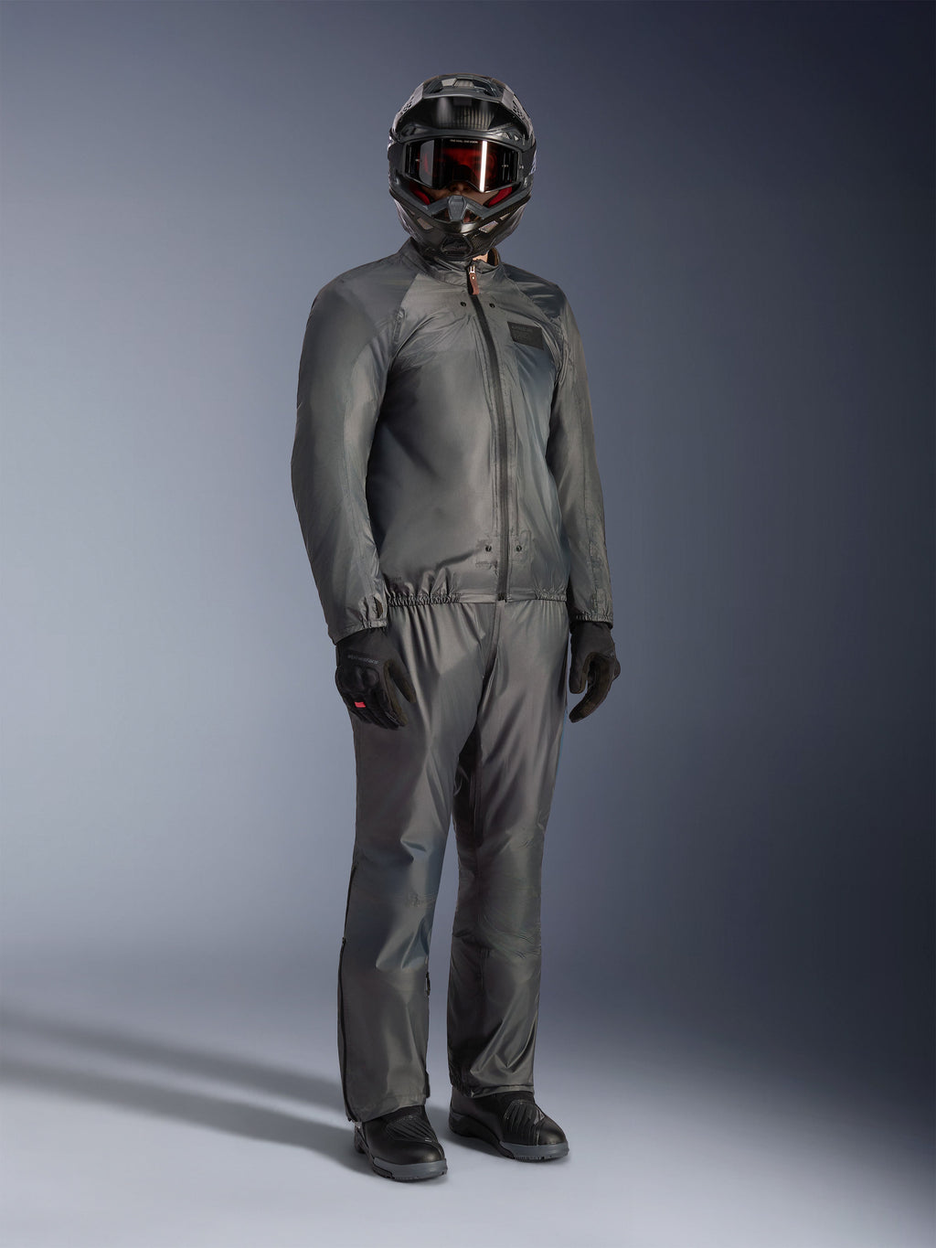 Motorrijder in de Alpinestars Borrego Drystar® Jas in Warm Gray, volledige adventure-uitrusting met bijpassende grijze broek, zwarte integraalhelm met motorbril en zwarte handschoenen, voorzien van een strak waterdicht textielontwerp.