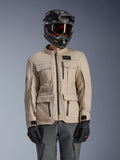 Alpinestars Borrego Drystar® jas, adventure motorjas, Warm Gray, met meerdere cargozakken aan de voorzijde en zwarte ritsen, gedragen met een zwarte helm, motorbril en donkergrijze motorbroek, robuust textielontwerp voor touring