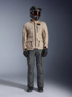 Alpinestars Borrego Drystar® Jas in Warm Gray, avontuurlijke motorjas met vier cargozakken aan de voorzijde, gecombineerd met een donkergrijze motorbroek, zwarte off-road helm en zwarte handschoenen, met een robuuste gewaxt canvas uitstraling