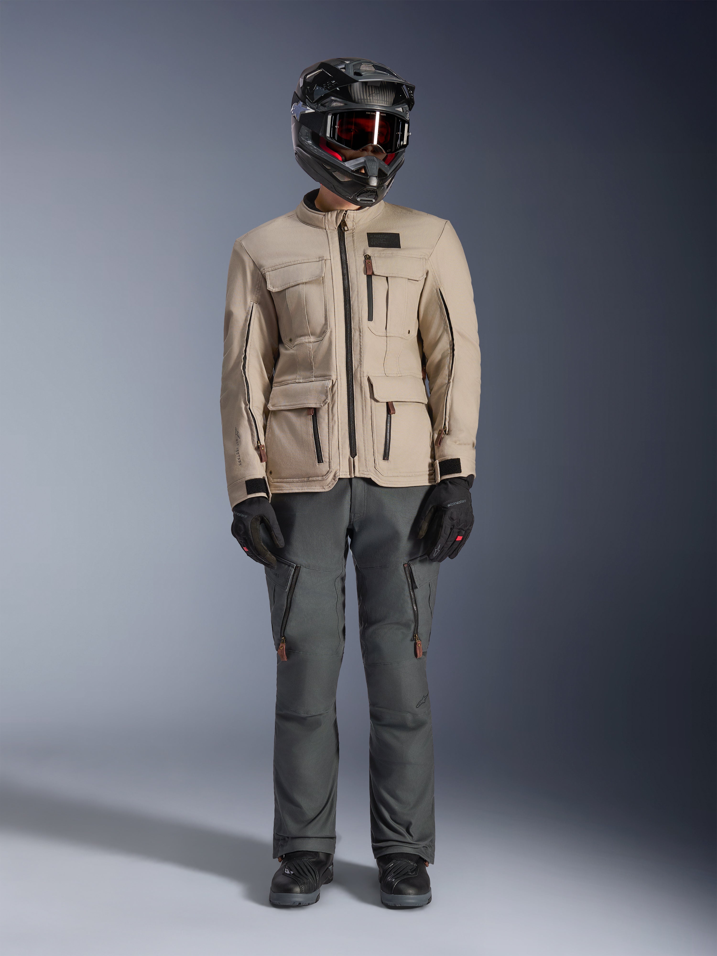 Alpinestars Borrego Drystar® Jas in Warm Gray, avontuurlijke motorjas met vier cargozakken aan de voorzijde, gecombineerd met een donkergrijze motorbroek, zwarte off-road helm en zwarte handschoenen, met een robuuste gewaxt canvas uitstraling