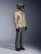 Motorrijder die de Alpinestars Borrego Drystar® Jas draagt in Warm Gray, adventure motorjas met een gewaxte canvastextuur en functionele zakken, gecombineerd met een donkergrijze motorbroek, een carbon helm, zwarte handschoenen en zwarte laarzen, studioweergave ten voeten uit van een complete adventure touring outfit.