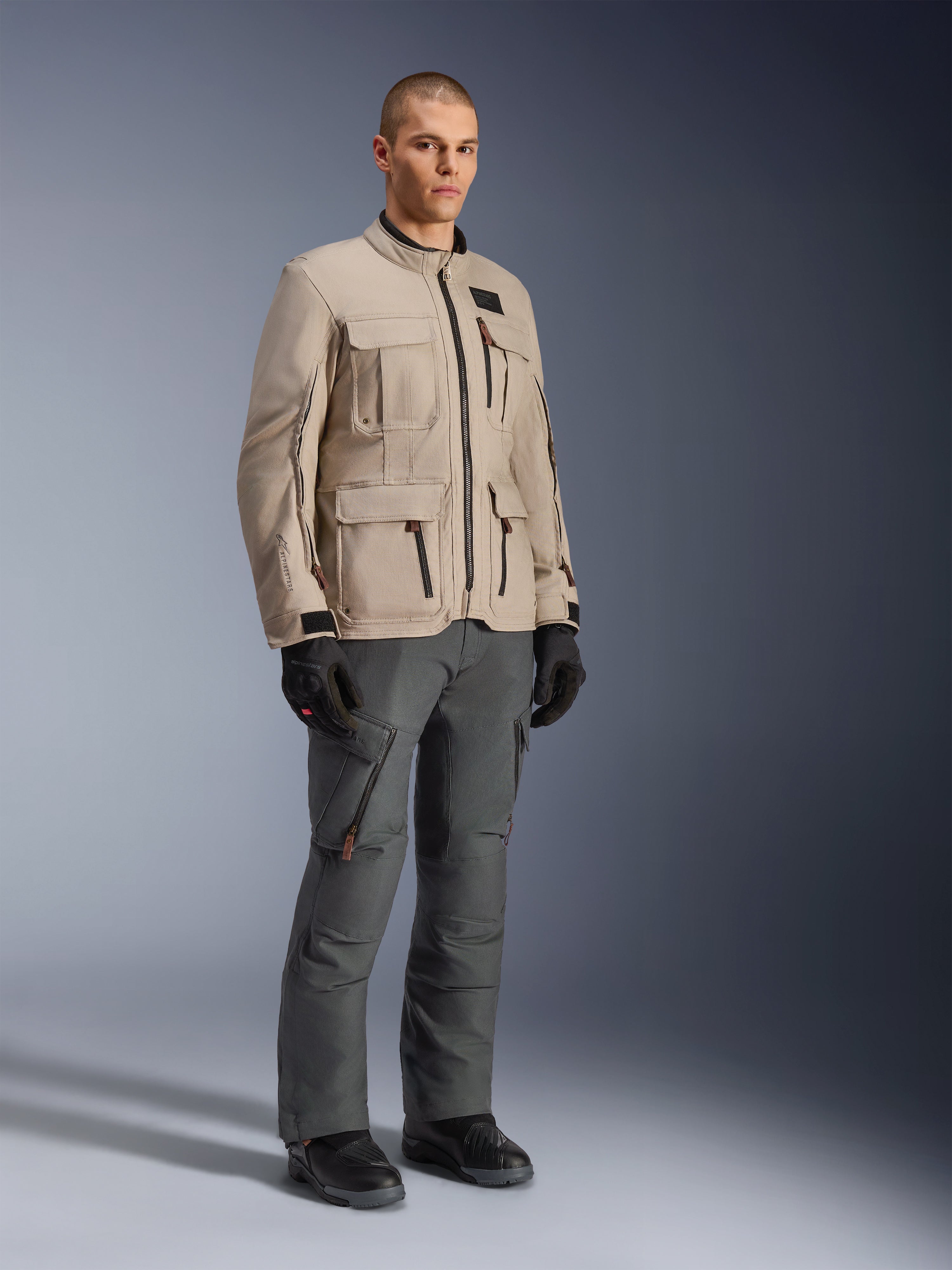 Alpinestars Borrego Drystar® jas in Warm Gray, adventure motorjas met een gewaxte-canvas uitstraling en meerdere functionele zakken, gedragen door een model met donkergrijze motorbroek, zwarte handschoenen en zwarte laarzen, full-body studioportret