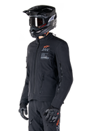 Alpinestars AMT-8 Stretch Drystar® XF Jas, zwart, gedragen door een motorrijder met bijpassende adventure broek en handschoenen, voorzien van een zwarte motorcrosshelm en bril, ventilatieopeningen met rits op de borst en mouwen, Alpinestars Motion Technology branding met een oranje logo, premium adventure-touring uitrusting