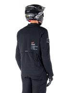 Achteraanzicht van een motorrijder die de zwarte Alpinestars AMT-8 Stretch Drystar® XF Jas draagt, voorzien van 'AMT Alpinestars Motion Technology' branding op de rug, technische ventilatie met ritssluiting, gecombineerd met een zwarte motorhelm en handschoenen
