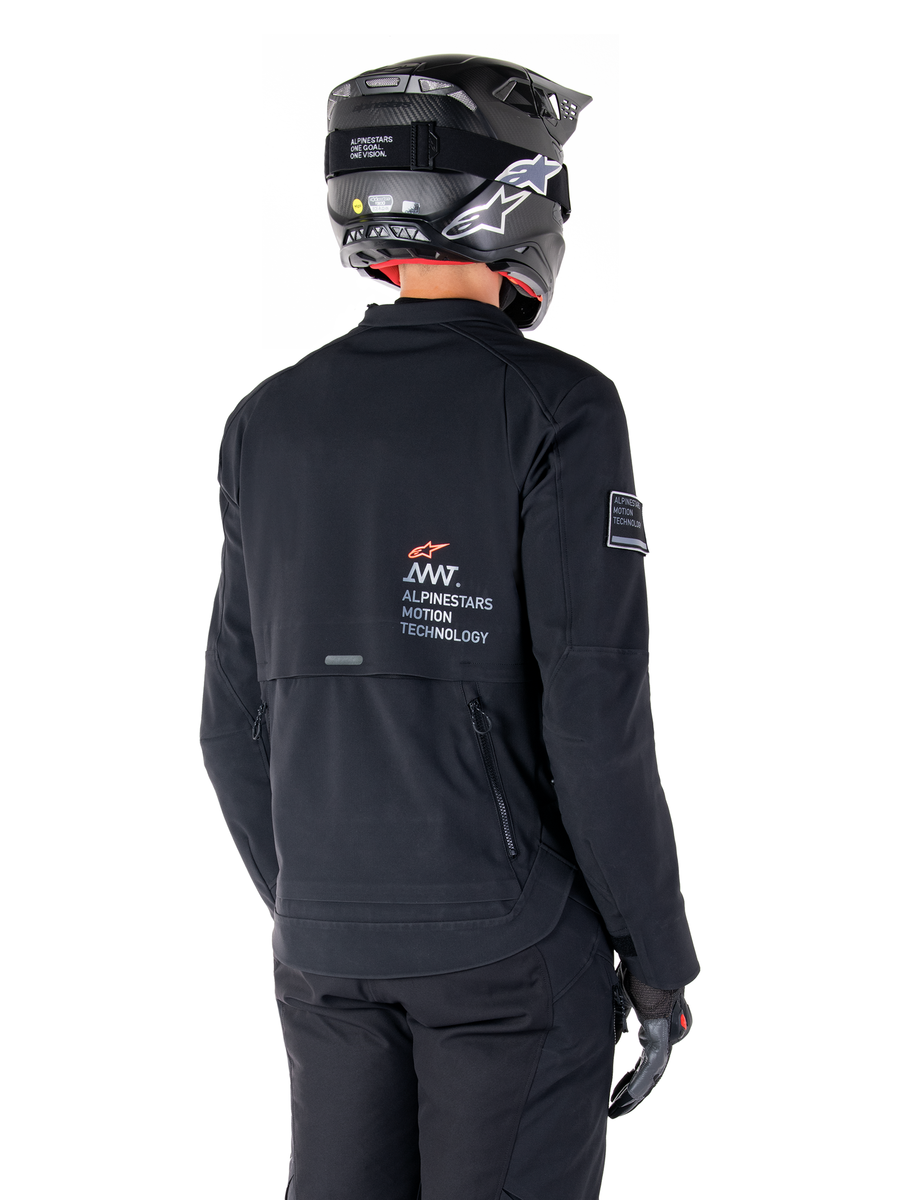 Achteraanzicht van een motorrijder die de zwarte Alpinestars AMT-8 Stretch Drystar® XF Jas draagt, voorzien van 'AMT Alpinestars Motion Technology' branding op de rug, technische ventilatie met ritssluiting, gecombineerd met een zwarte motorhelm en handschoenen