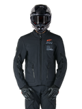 Alpinestars AMT-8 Stretch Drystar® XF Jas, zwarte motorjas, voorzien van een oranje Alpinestars-logo en grijze AMT-branding, hoge kraag met rits aan de voorzijde en ventilatieopeningen, gedragen met zwarte helm en handschoenen, beschermende adventure touring-uitrusting