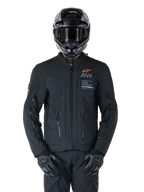 Alpinestars AMT-8 Stretch Drystar® XF Jas, zwarte motorjas, voorzien van een oranje Alpinestars-logo en grijze AMT-branding, hoge kraag met rits aan de voorzijde en ventilatieopeningen, gedragen met zwarte helm en handschoenen, beschermende adventure touring-uitrusting