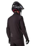 Alpinestars AMT-8 Stretch Drystar® XF voering, zwarte waterdichte voering voor motorjas, achteraanzicht van een rijder met zwarte helm en handschoenen, voorzien van Alpinestars Motion Technology-branding op de onderrug, minimalistisch en functioneel ontwerp voor adventure ritten.