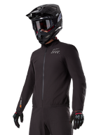Rijder draagt de Alpinestars AMT-8 Stretch Drystar® XF Liner, zwarte wind- en waterdichte stretchjas, met AMT-logo op de borst en Drystar XF-branding op de mouw, gecombineerd met een zwarte adventure-helm en beschermende handschoenen.